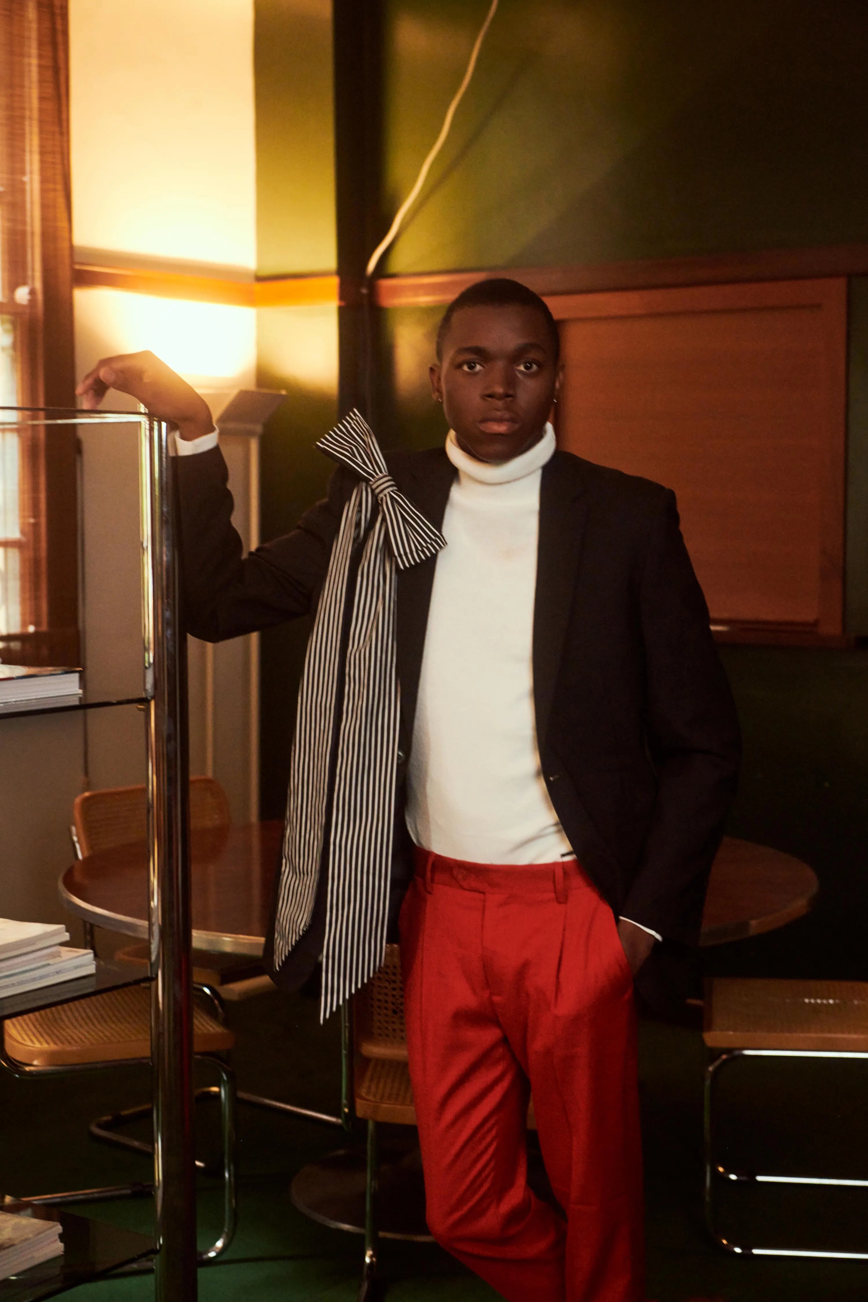 A man wearing a black blazer, white turtleneck, red trousers, and a striped bow brooch, standing in a dimly lit room with warm lighting.