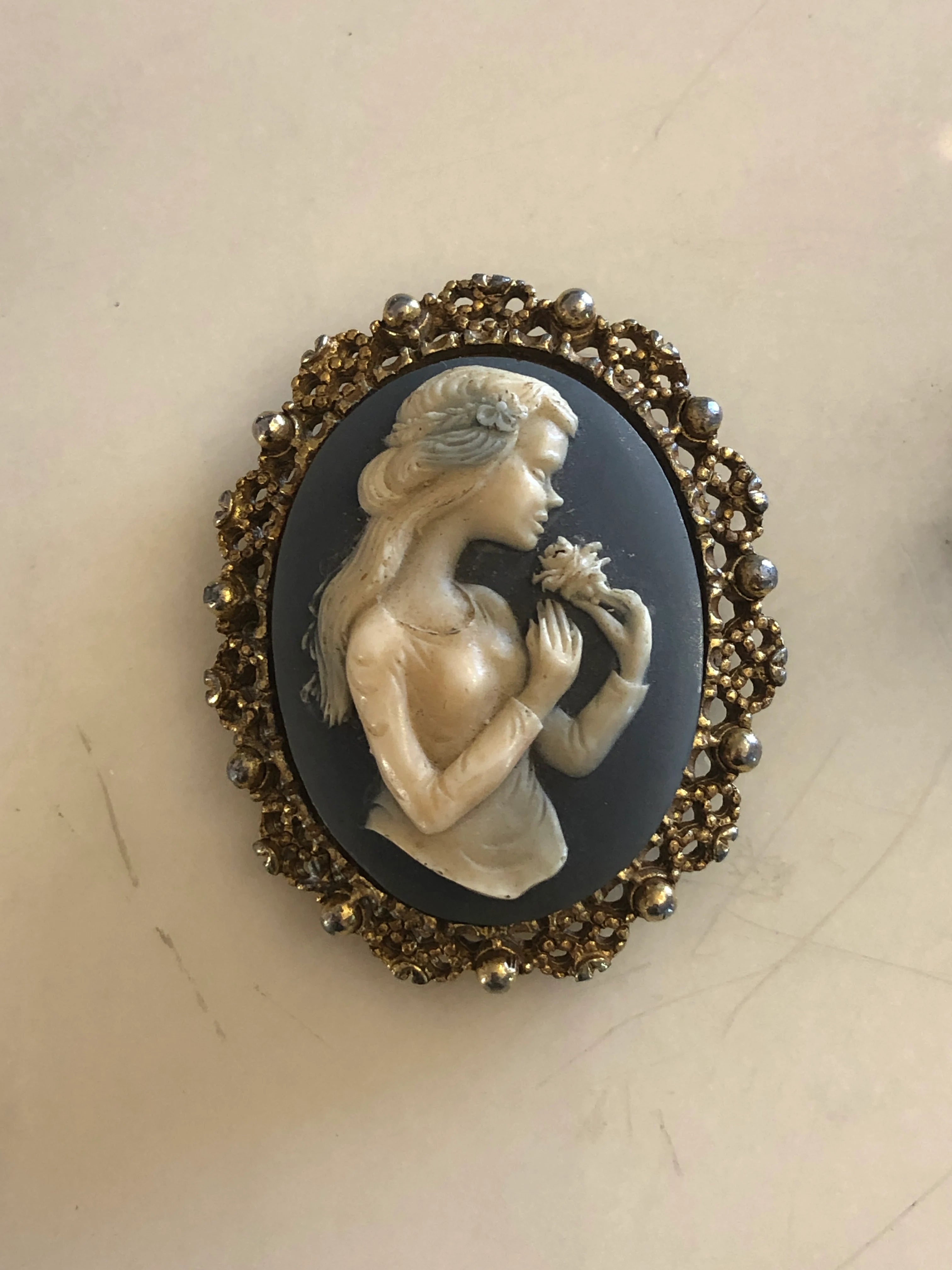 Broochella bleu vintage cameo brooch. It features a gold decorative frame and a blue and white cameo with the profile of a woman with long hair holding a flower. The background is white marble