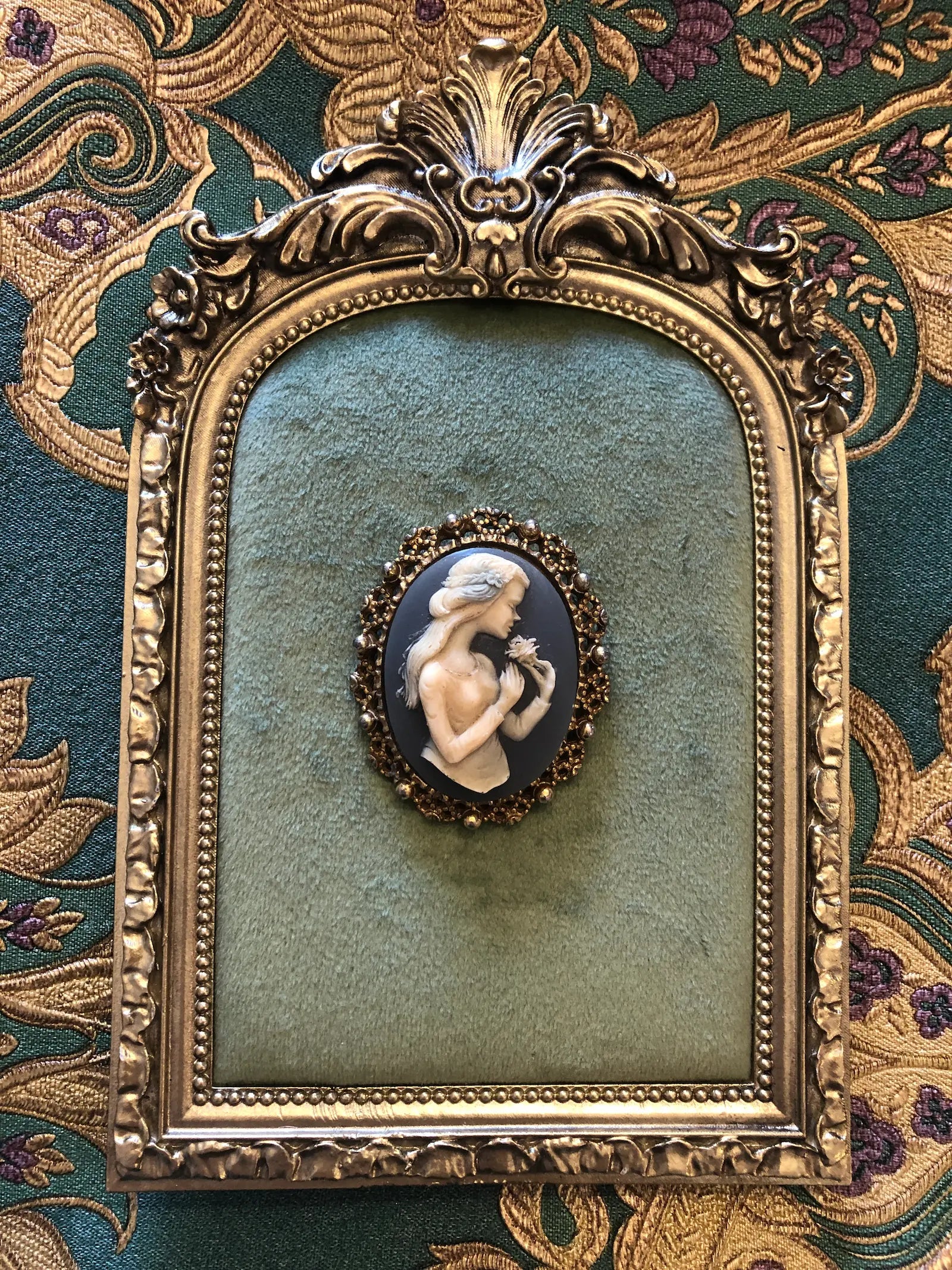 Broochella bleu vintage cameo brooch. It features a gold decorative frame and a blue and white cameo with the profile of a woman with long hair holding a flower. It is pinned to a green velvet tray with an ornate gold frame. The background is green and gold damask wallpaper.