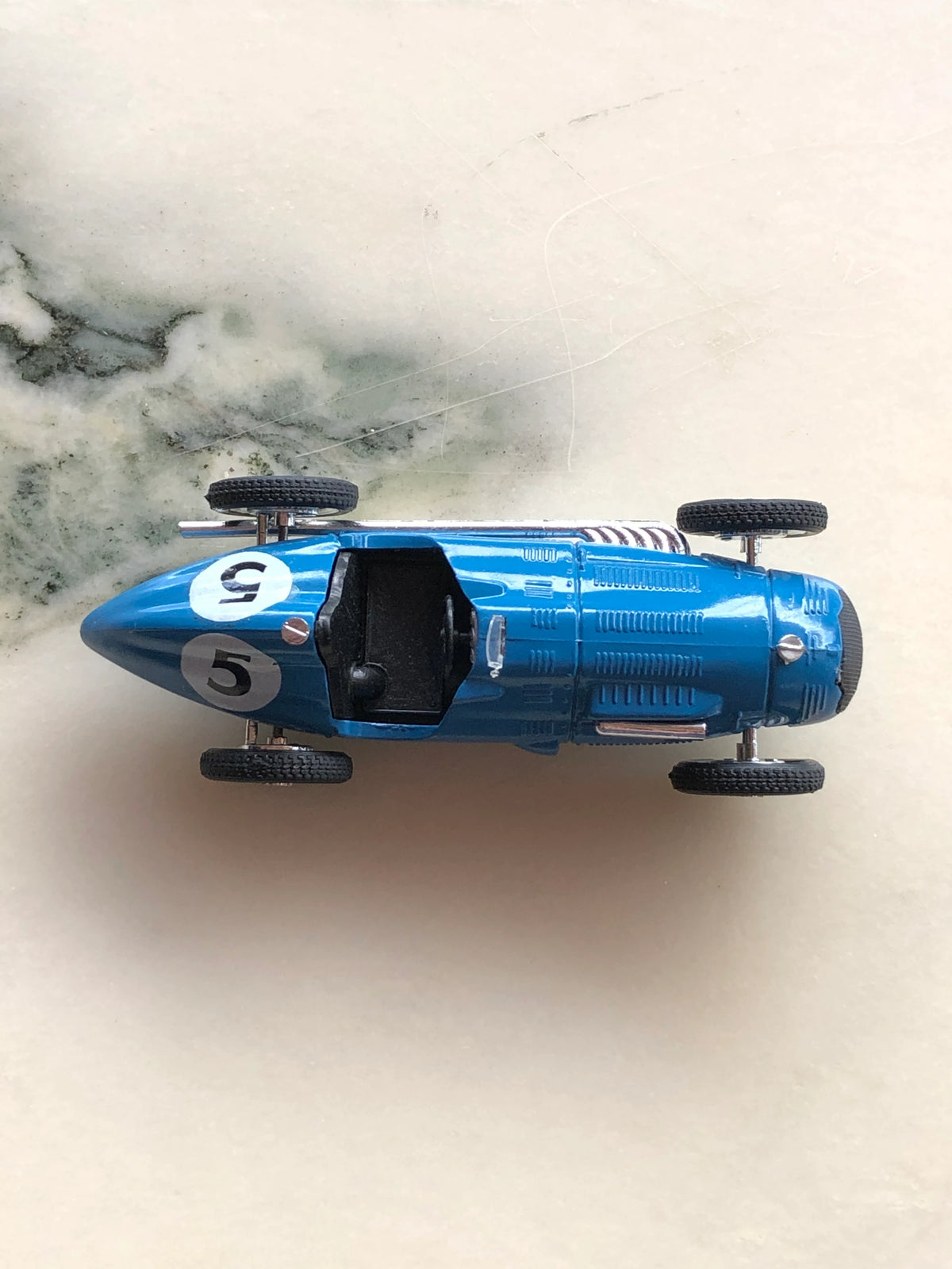 Broochella Blu Veloce No.5 Vintage Grand Prix Racing Car Brooch photographed from above, showcasing its streamlined blue body, black open cockpit, white number 5 racing decals, silver engine detailing and four black tyres.