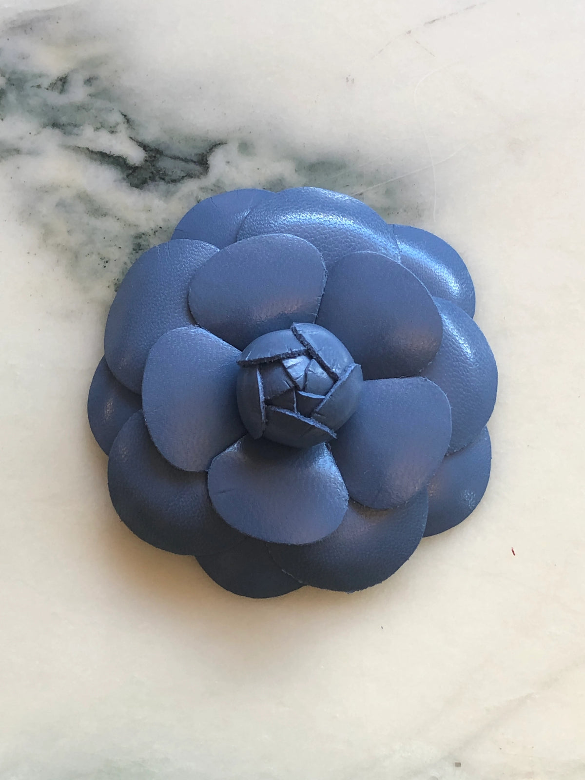 A Broochella Camelia handmade blue leather flower-shaped accessory brooch,  resting on a white marble surface.