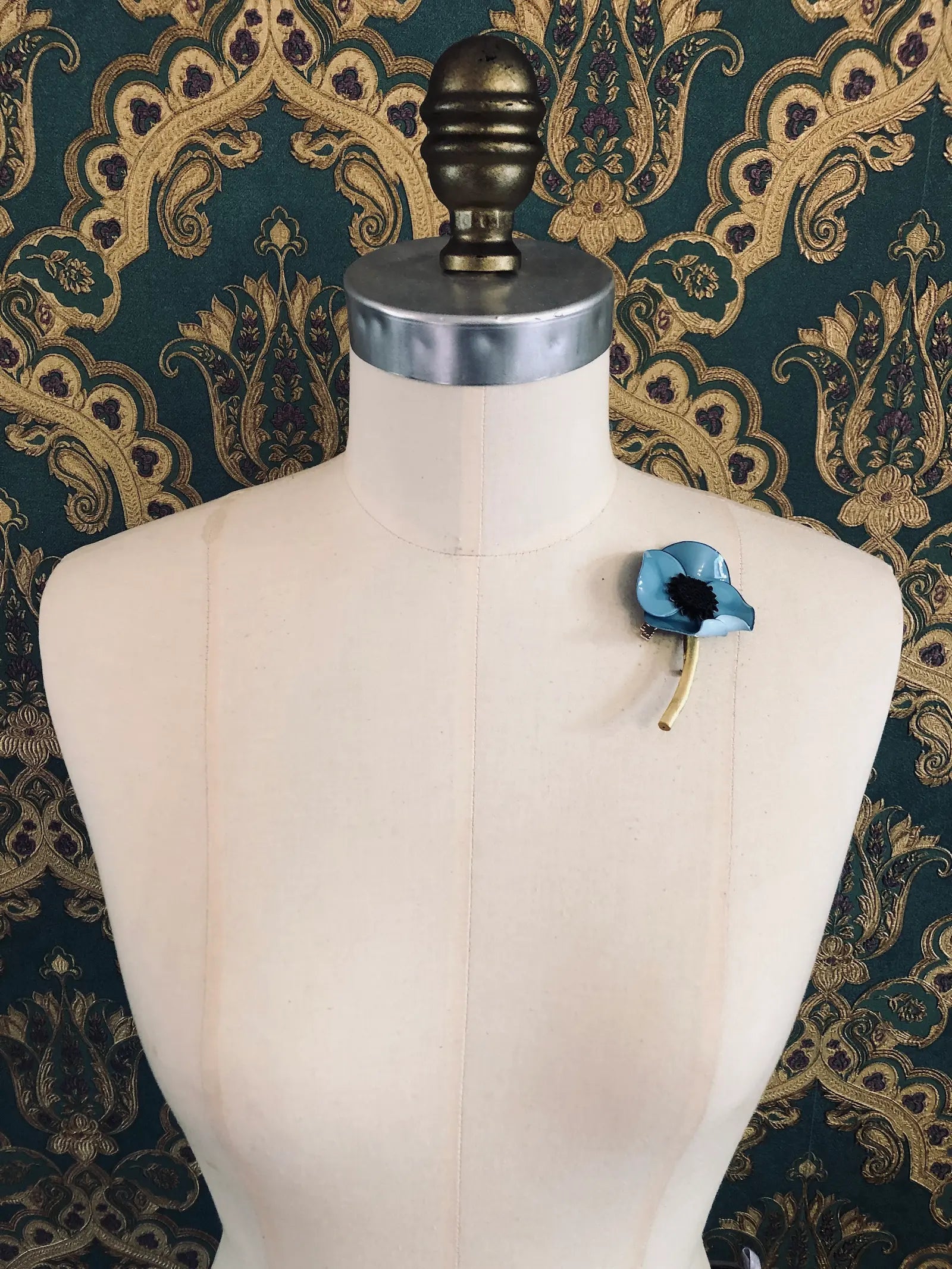 Broochella x Fabrice Paris red moulded acetate poppy flower brooch in blue. It is pinned to a mannequin. The background is green and gold damask wallpaper. 