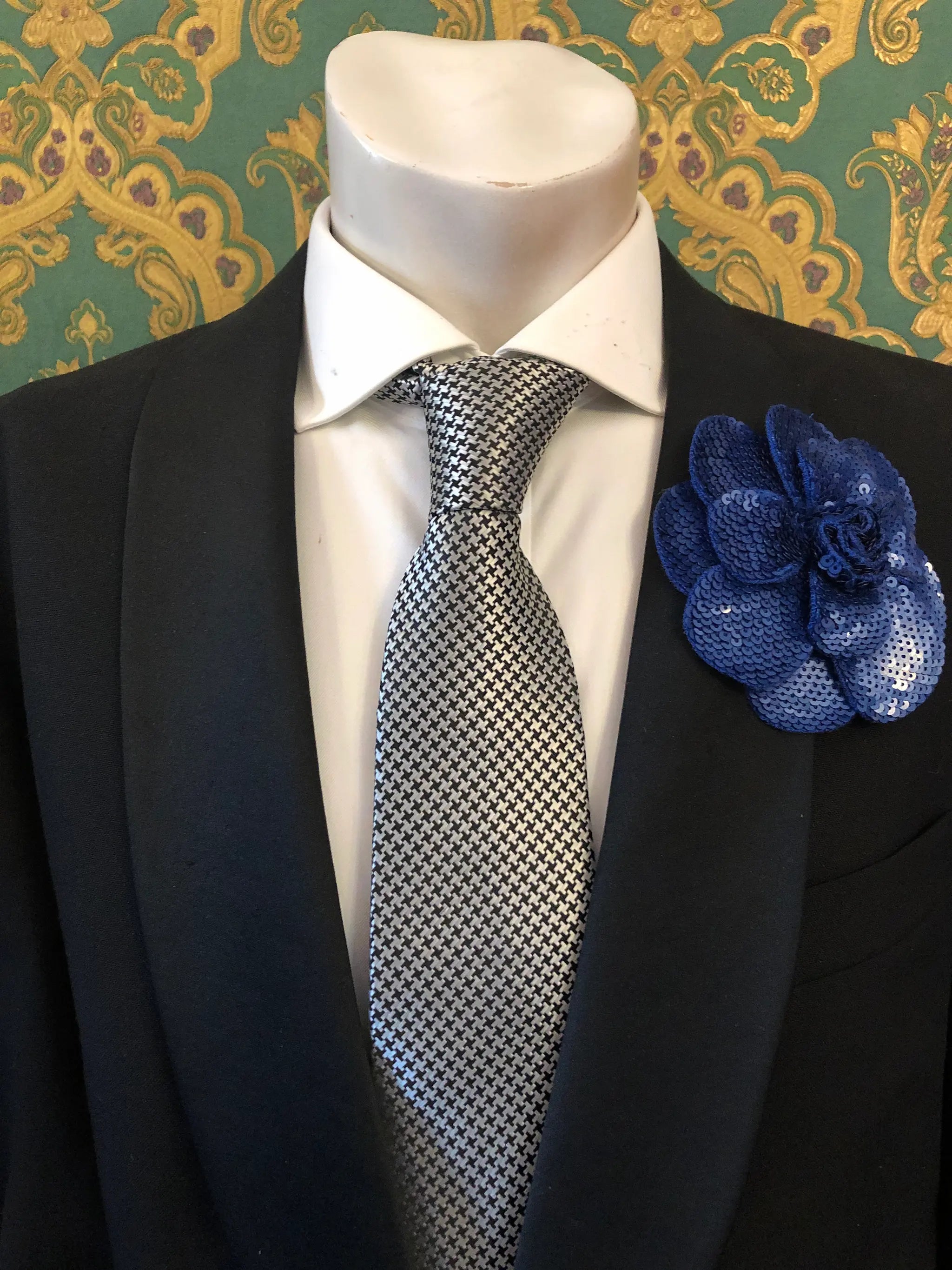 A men's formal accessory set displayed on a white mannequin torso, featuring a black suit jacket, a white dress shirt, a silver and black checkered tie, and an orange Men's Broochella Scintillante sequinned flower brooch lapel pin against a green and gold patterned wallpaper background.