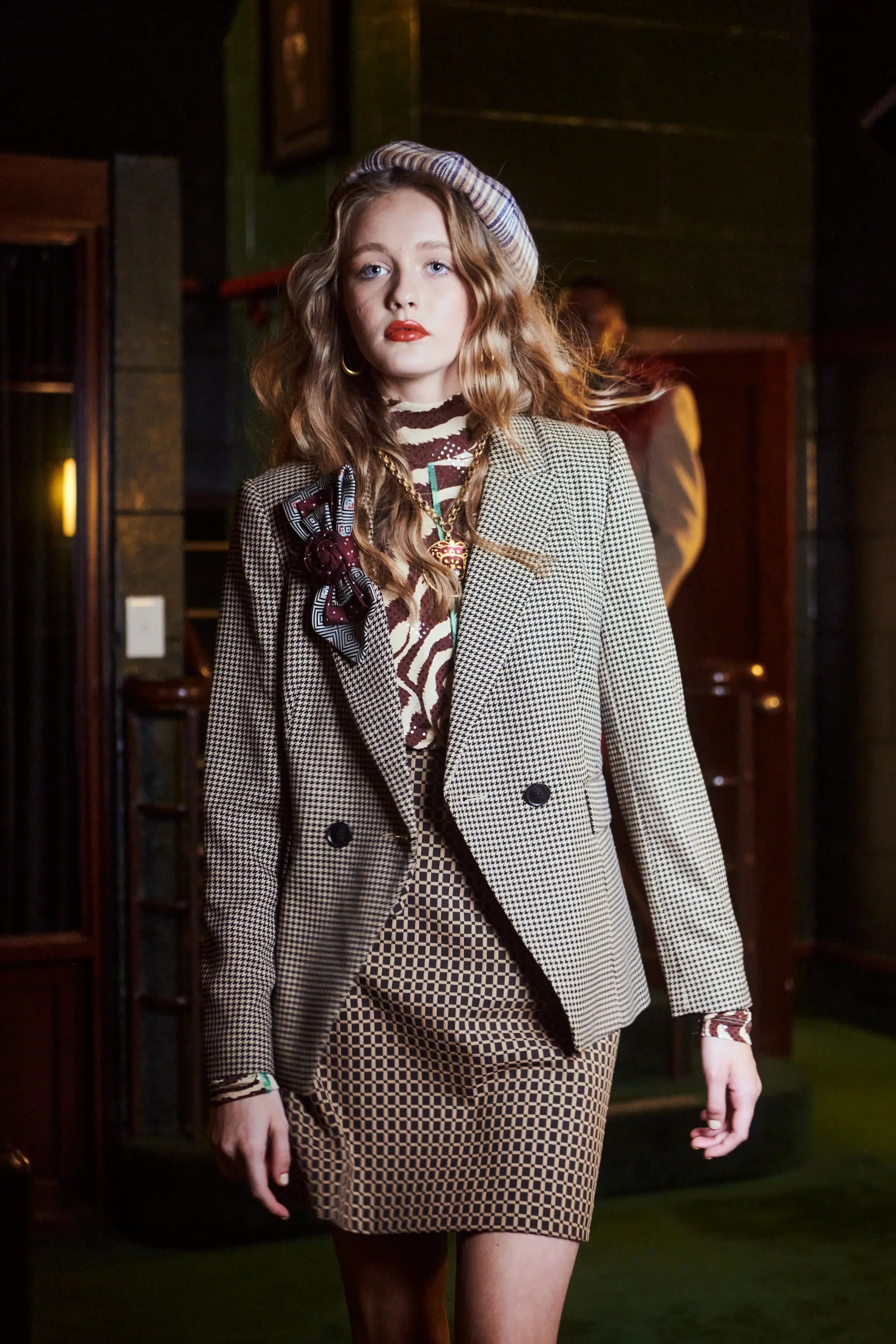 A woman models a vintage-style double-breasted blazer in a beige and brown houndstooth pattern, paired with a matching patterned mini skirt, a patterned scarf, and a striped beret, set against a dark, atmospheric background. She is wearing the Broochelal bolivar cravatte tie brooch