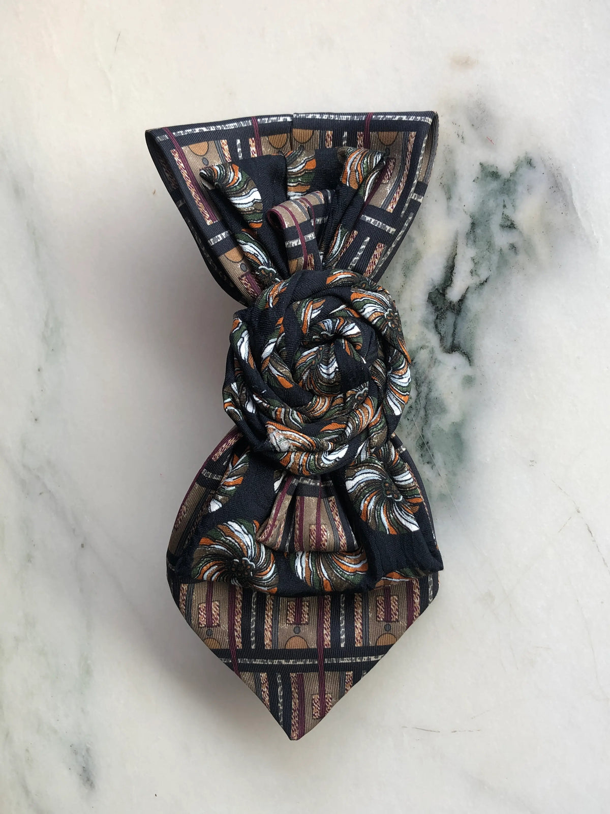 Broochella Bolivar Cravatte Tie Brooch Pin No. 1, a one-of-a-kind statement brooch handmade from repurposed vintage patterned ties in black, brown, burgundy, cream and rust, with a sculptural rosette centre, photographed on white marble with grey-green veining.