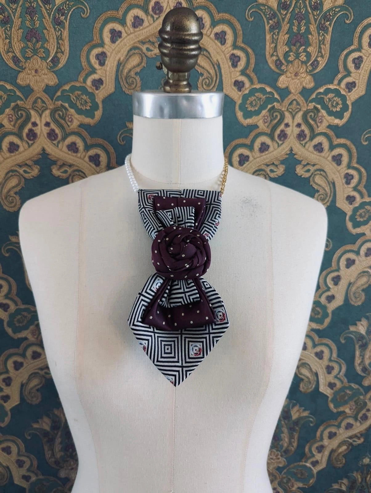 Broochella Bolivar Cravatte hand made up cycled vintage tie brooch in maroon-number 10. it is pinned to a mannequin. The background is green and gold damask wallpaper