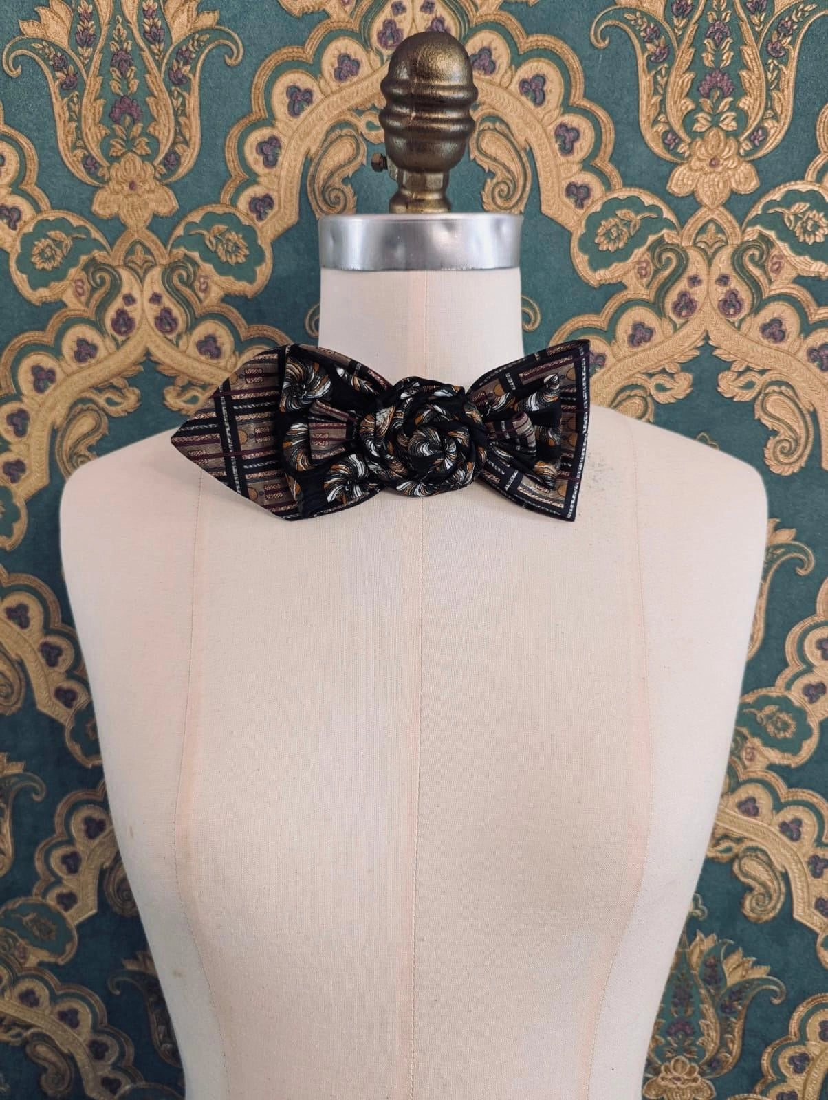 A Broochella Bolivar vintage upcycled necktie styled on a cream fabric dress form mannequin, featuring a black and brown geometric pattern with a central fabric rose embellishment. The background is green and gold damask wallpaper.