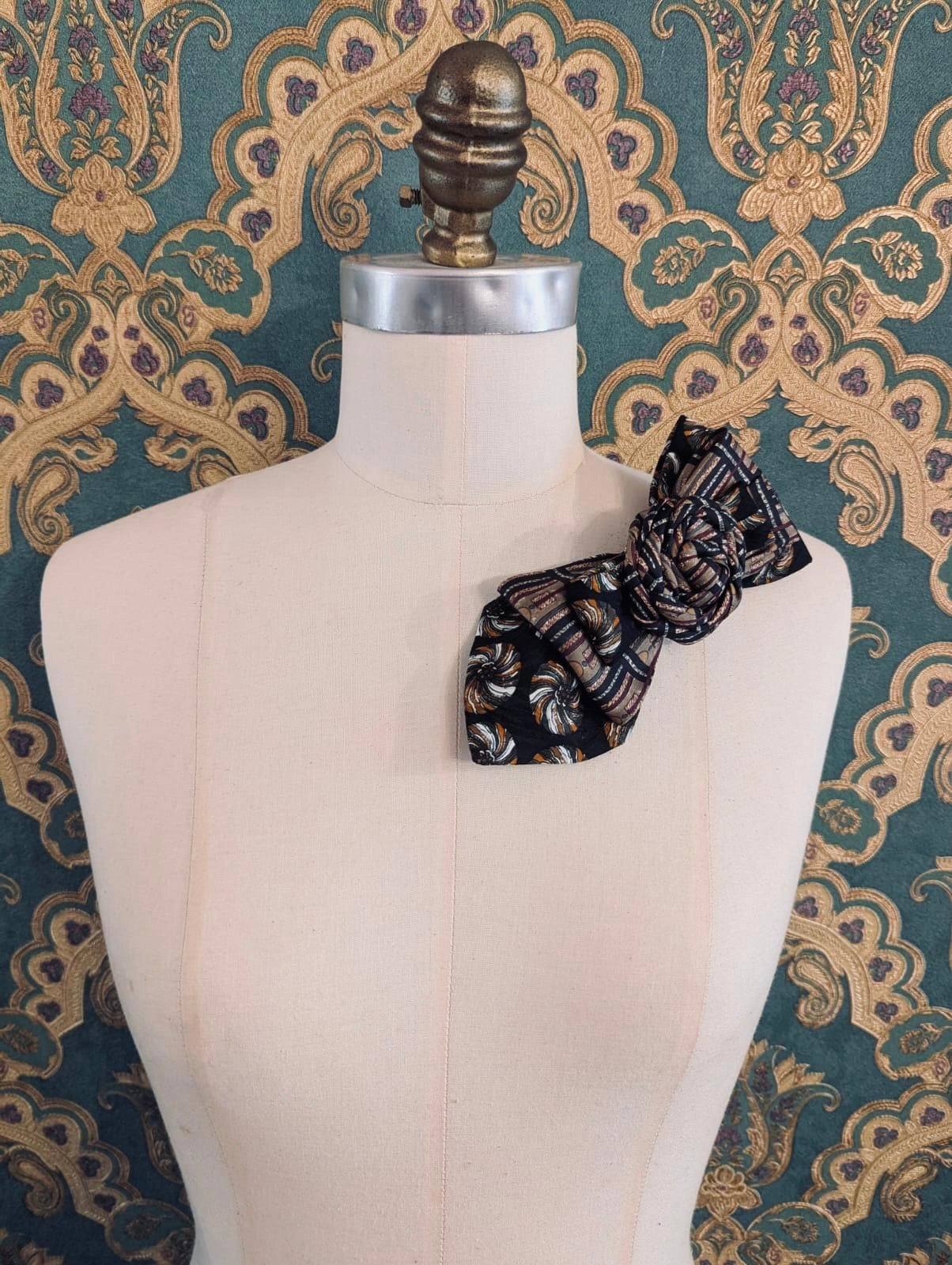 A Broochella Bolivar vintage upcycled necktie styled on a cream fabric dress form mannequin, featuring a black and brown geometric pattern with a central fabric rose embellishment. The background is green and gold damask wallpaper.