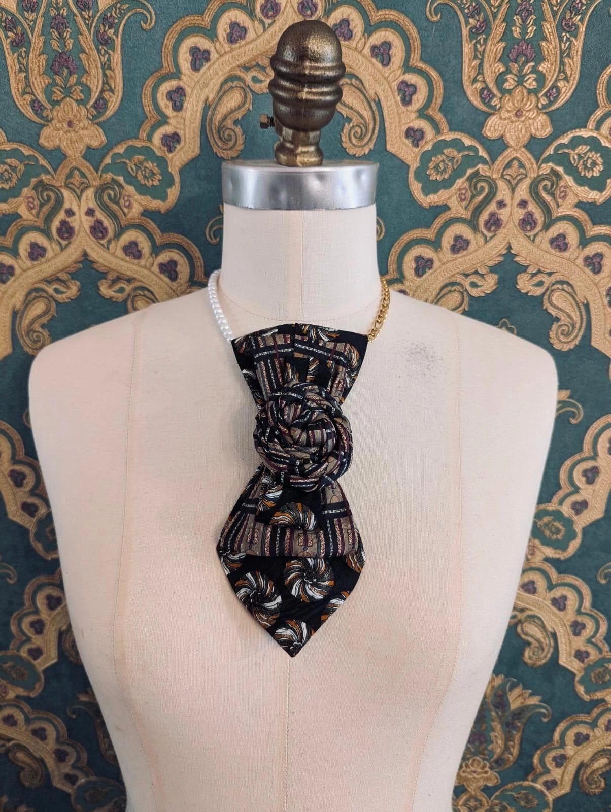 A Broochella Bolivar vintage upcycled necktie styled on a cream fabric dress form mannequin, featuring a black and brown geometric pattern with a central fabric rose embellishment. The background is green and gold damask wallpaper.