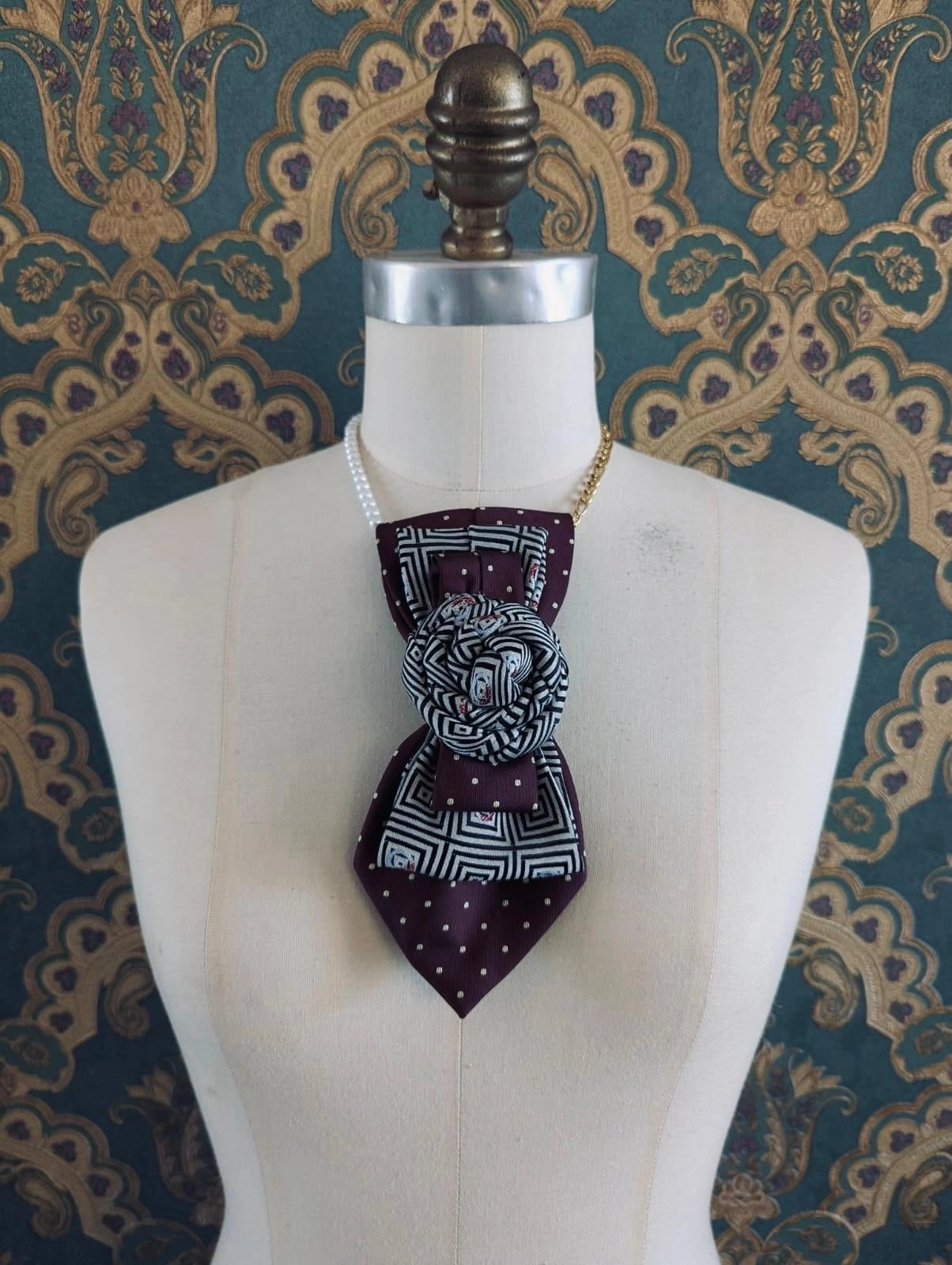 A Broochella Bolivar vintage upcycled necktie styled on a cream fabric dress form mannequin, featuring a burgundy and white geometric pattern with a central fabric rose embellishment and a gold chain strap. The background is green and gold damask wallpaper.