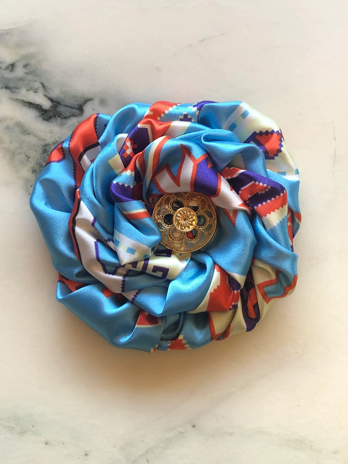 Broochella Bougainville Flower Brooch featuring sculptural blue printed satin petals with red, purple and ivory detailing and an ornate gold-tone centre, photographed on cream marble.