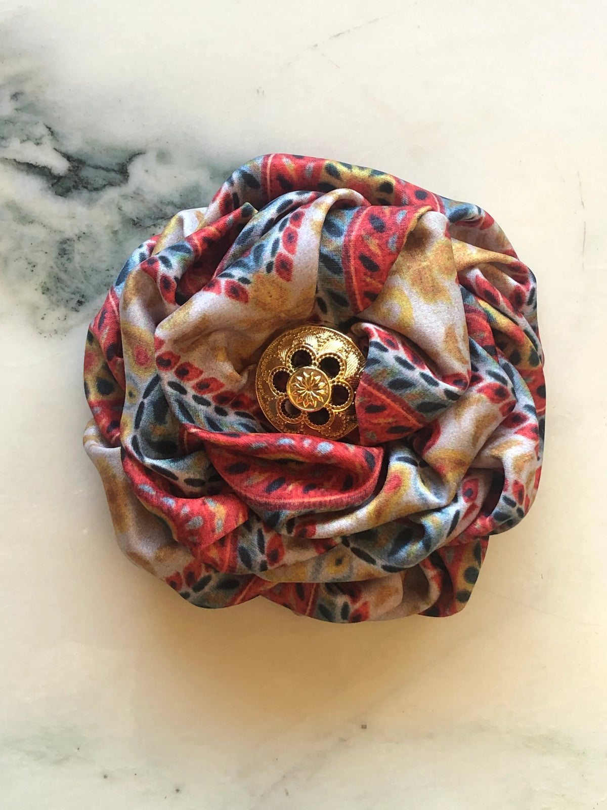 Broochella Bougainville Flower Brooch featuring sculptural printed satin petals in orange, red, gold and blue tones with an ornate gold-tone centre, photographed on cream marble.