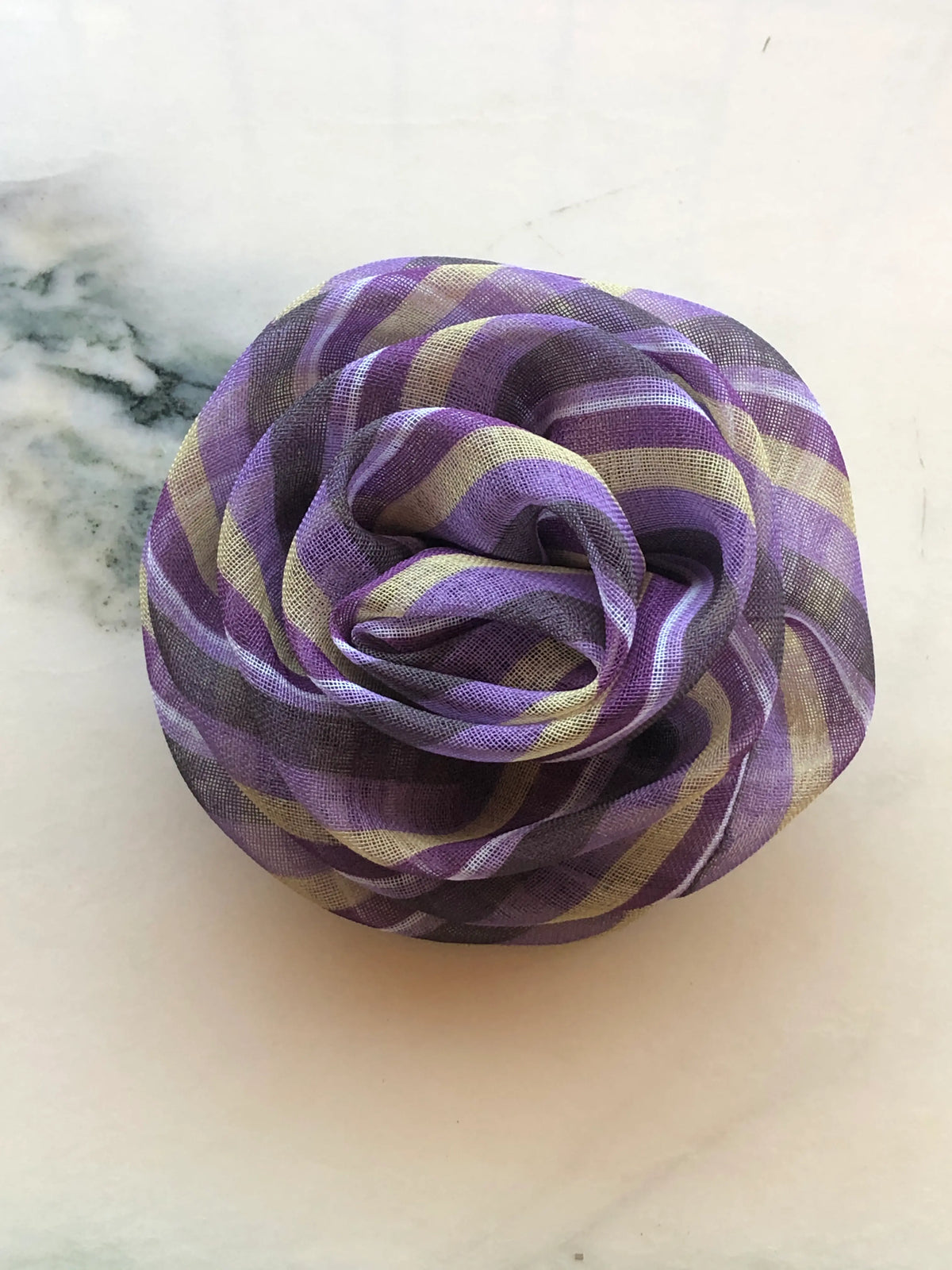 Broochella Brassica Flower Brooch in purple, featuring sculptural sheer fabric petals with a striped print in purple, lilac, cream and charcoal, arranged in a full rose-like design on a cream marble background.