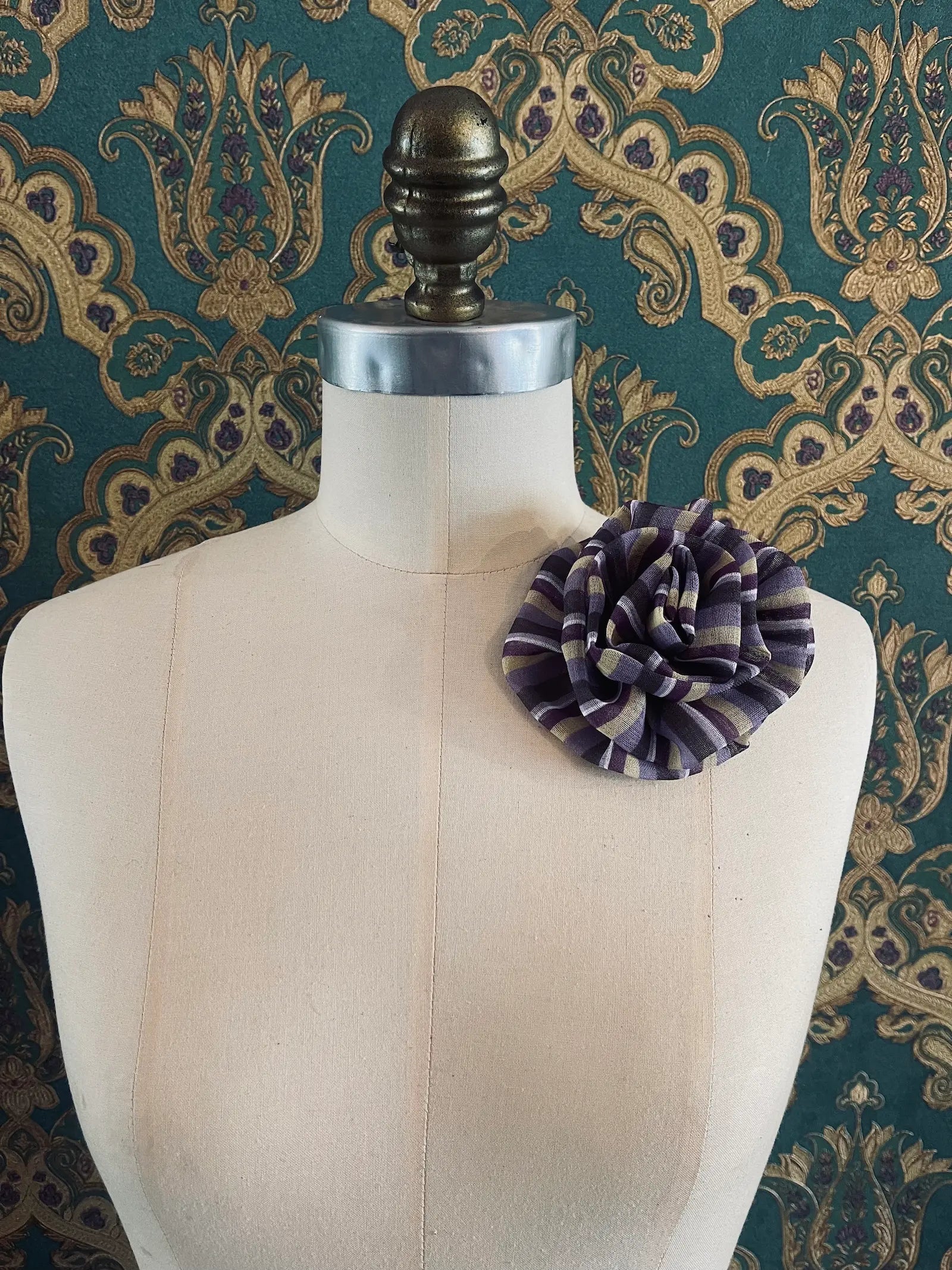 Broochella brassica handmade fabric flower brooch in purple and green stripes. Styled on a mannequin. The background is green and gold damask wallpaper