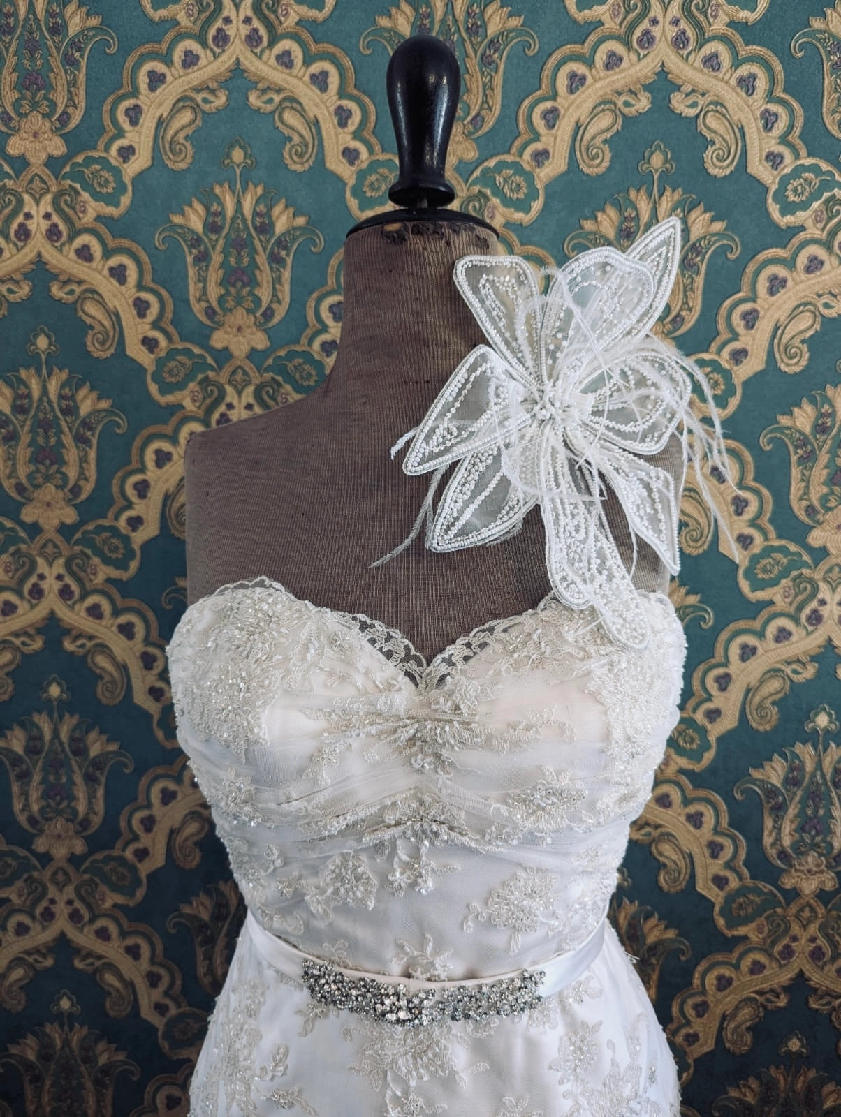 Broochella bridal Azlea hand beaded, white wedding flower brooch. Pictured on a mannequin that is wearing a white, beaded strapless wedding dress. The background is green and gold damask wallpaper. 