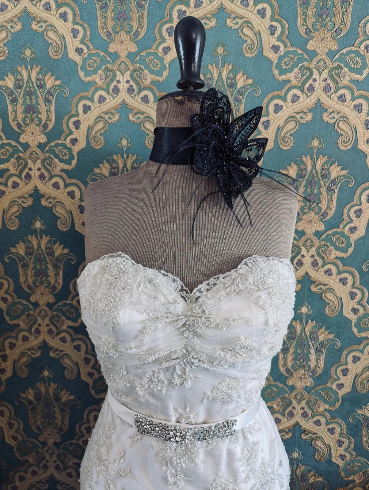 Broochella azalea small bridal, hand beaded, black, large flower brooch, with lace and crystal brooch embellishment. Pinned on a mannequin. The mannequin is dressed in a white, beaded, strapless wedding dress. The background is green and gold damask wallpaper.