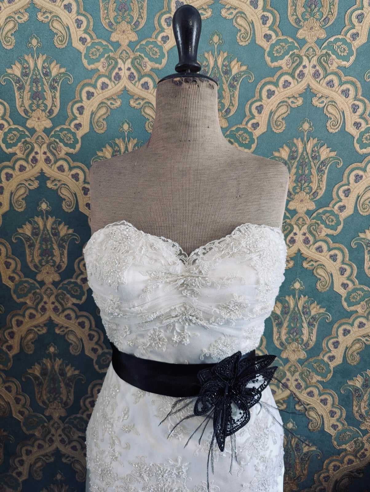 Broochella azalea small bridal, hand beaded, black, large flower brooch, with lace and crystal brooch embellishment. Pinned on a mannequin. The mannequin is dressed in a white, beaded, strapless wedding dress. The background is green and gold damask wallpaper.