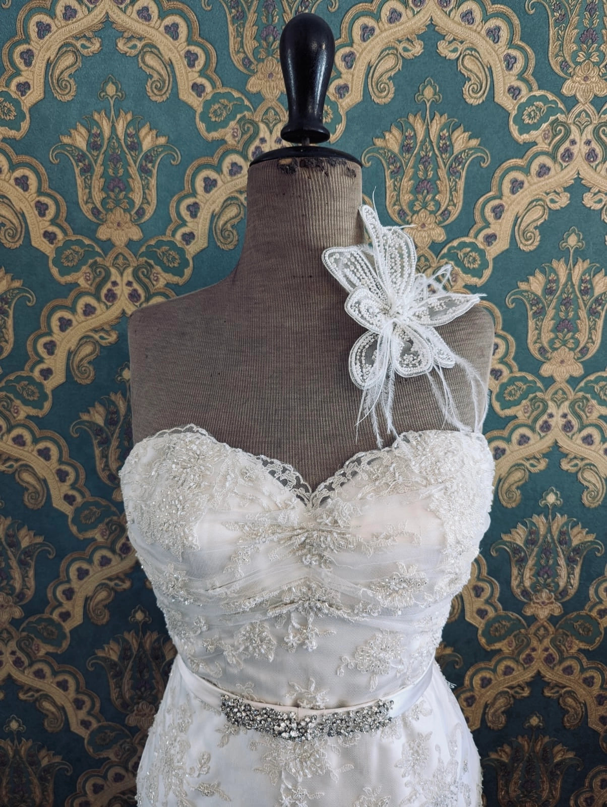 Broochella bridal Azlea hand beaded, small, white wedding flower brooch. Pictured on a mannequin that is wearing a white, beaded strapless wedding dress. The background is green and gold damask wallpaper. 