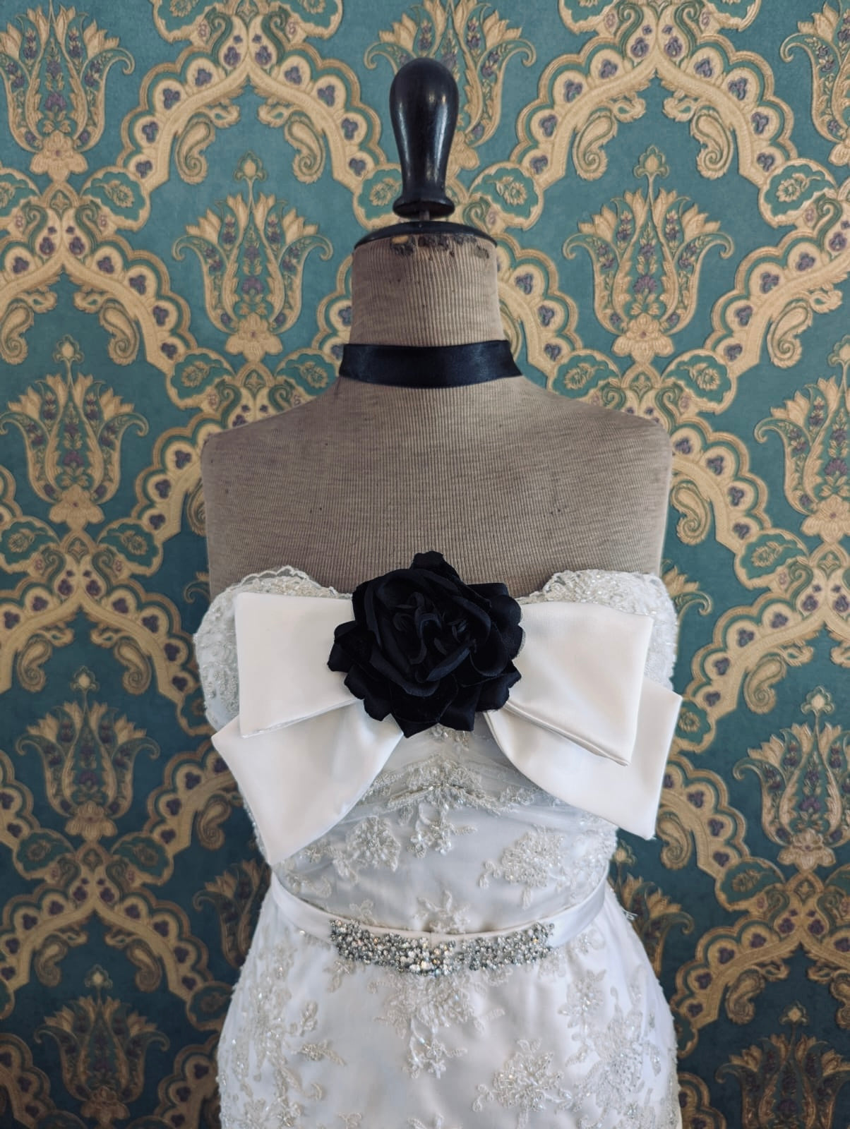 Broochella  dolce amore handmade bridal brow brooch in ivory silk satin. There is a black fabric flower embellishment at the centre of the bow. Pictured on a mannequin that is wearing a white, beaded strapless wedding dress. The background is green and gold damask wallpaper. 