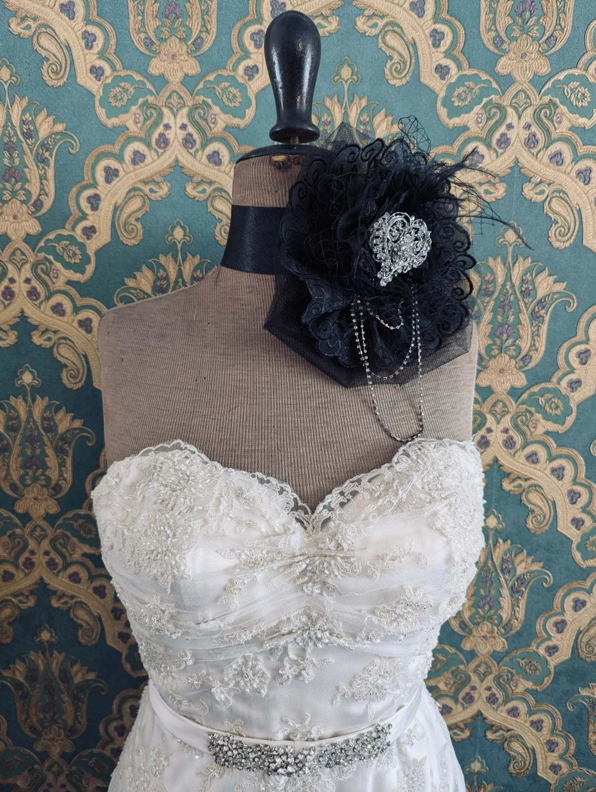 Broochella madonna bridal, hand beaded, black, large flower brooch, with lace and crystal brooch embellishment. Pinned on a mannequin. The mannequin is dressed in a white, beaded, strapless wedding dress. The background is green and gold damask wallpaper.