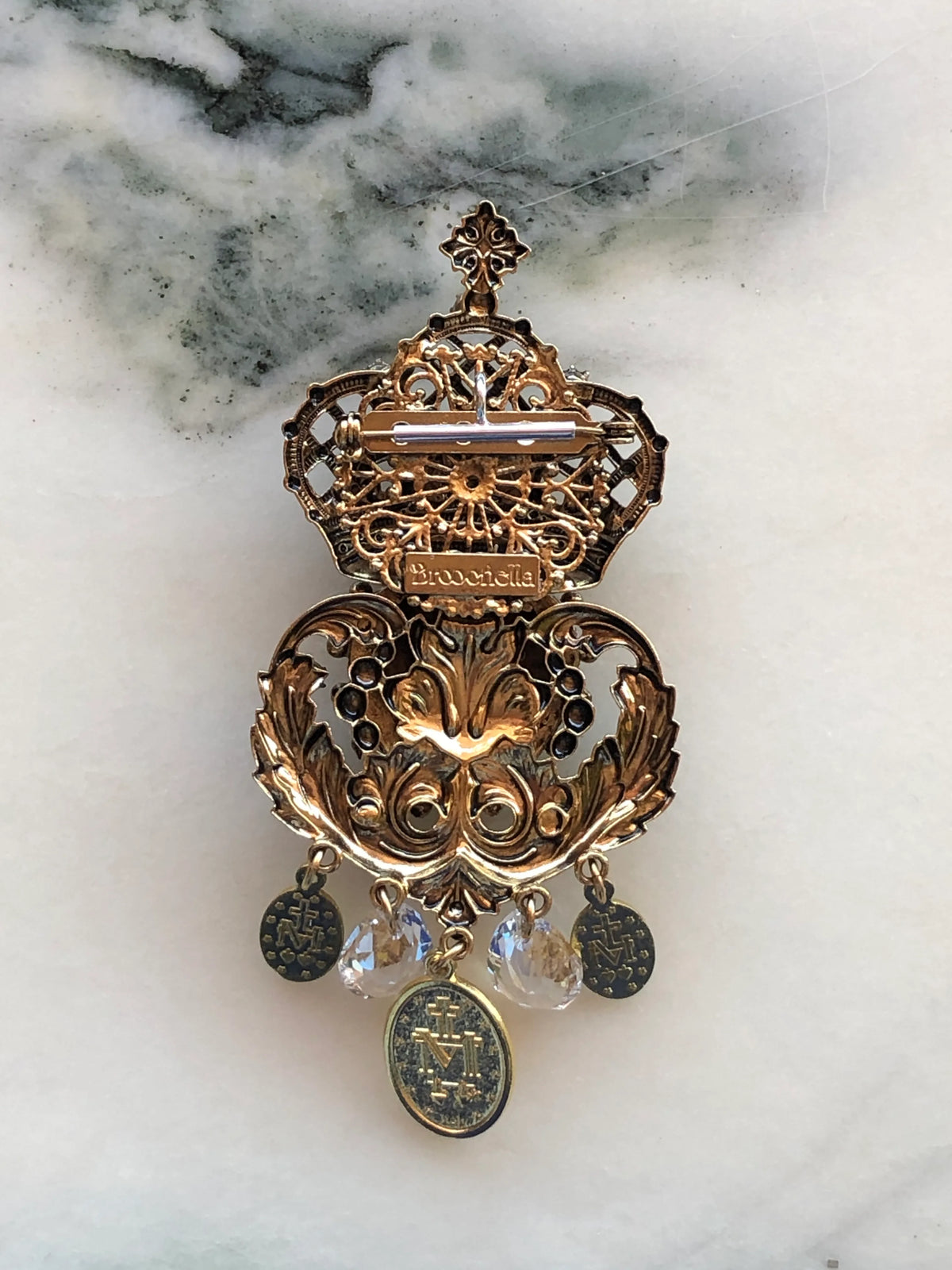 Broochella extended bar brooch to necklace converter in gold-tone metal with central oval loop. It is attached to a Broochella Agatha Sacro Cuore brooch and a silver chain, demonstrating how it works. photographed on white marble with grey-green veining.