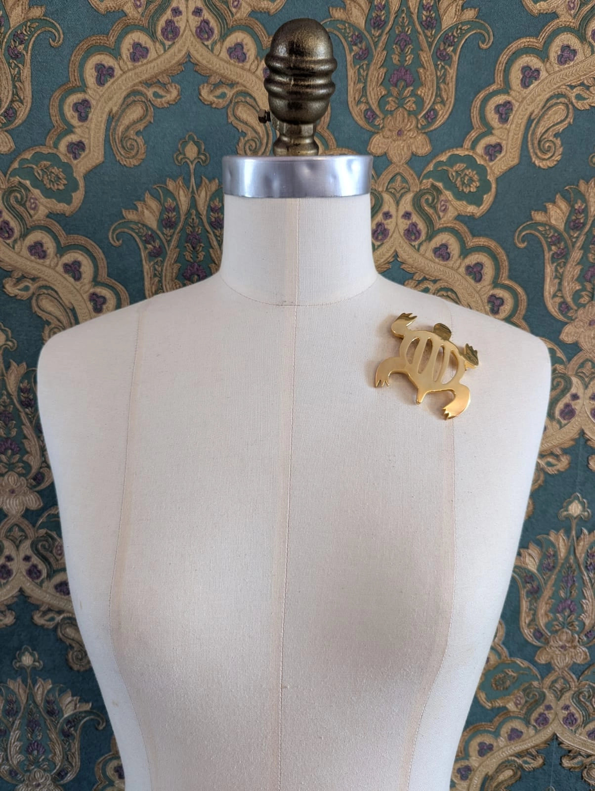 Broochella brooches with meaning designer gold metal brooch. Pictured on a mannequin. The background is green and gold damask wallpaper
