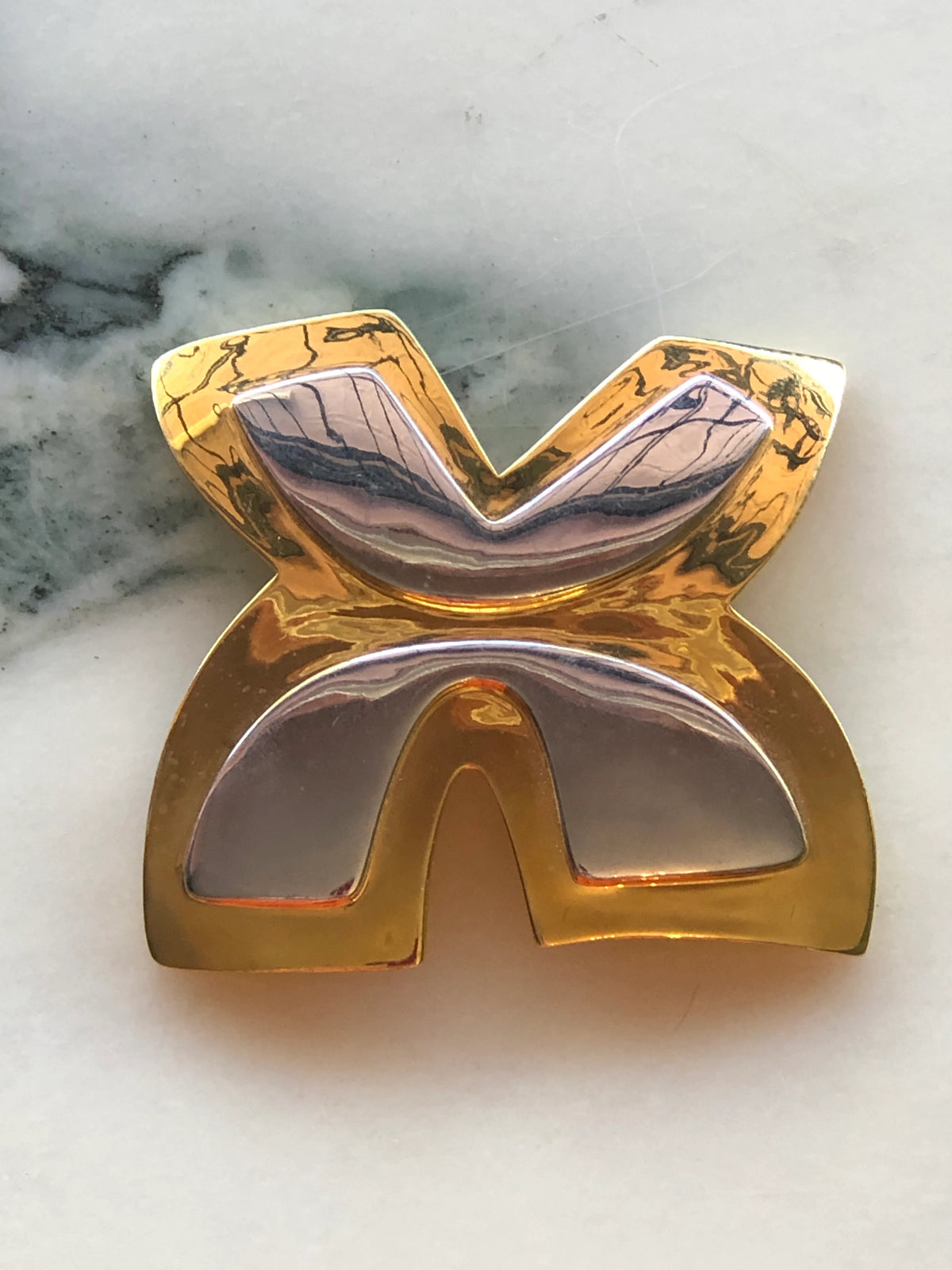 Broochella Brooches with Meaning Freedom Brooch Pin featuring a sculptural gold-tone geometric design with polished silver-tone accents on a cream marble background.