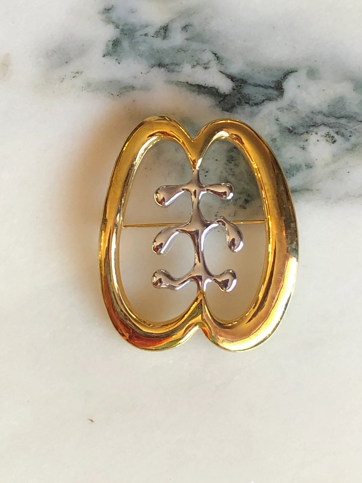 Broochella Brooches with Meaning – Friendship Brooch Pin, featuring a polished gold-tone oval design with an abstract silver-tone friendship symbol at the centre on a cream marble background.