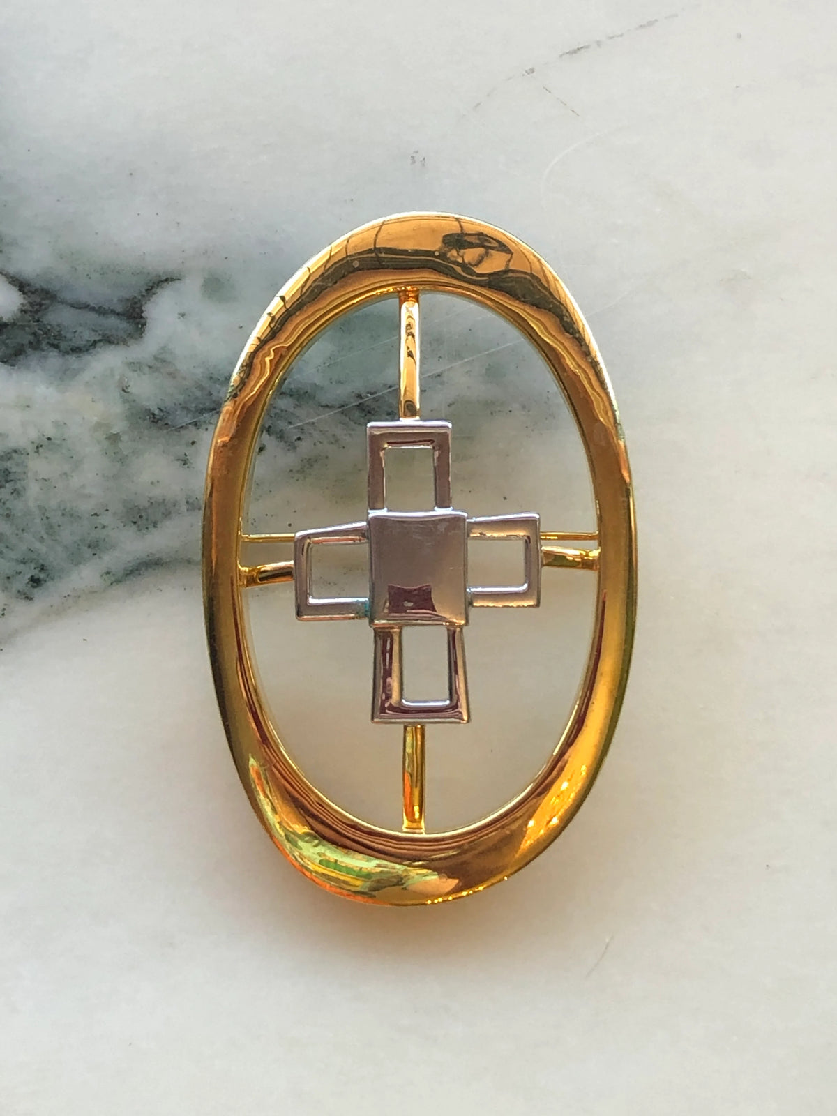 Broochella Brooches with Meaning Knowledge Brooch Pin featuring a polished gold-tone oval frame with a geometric silver-tone cross motif on a cream marble background.