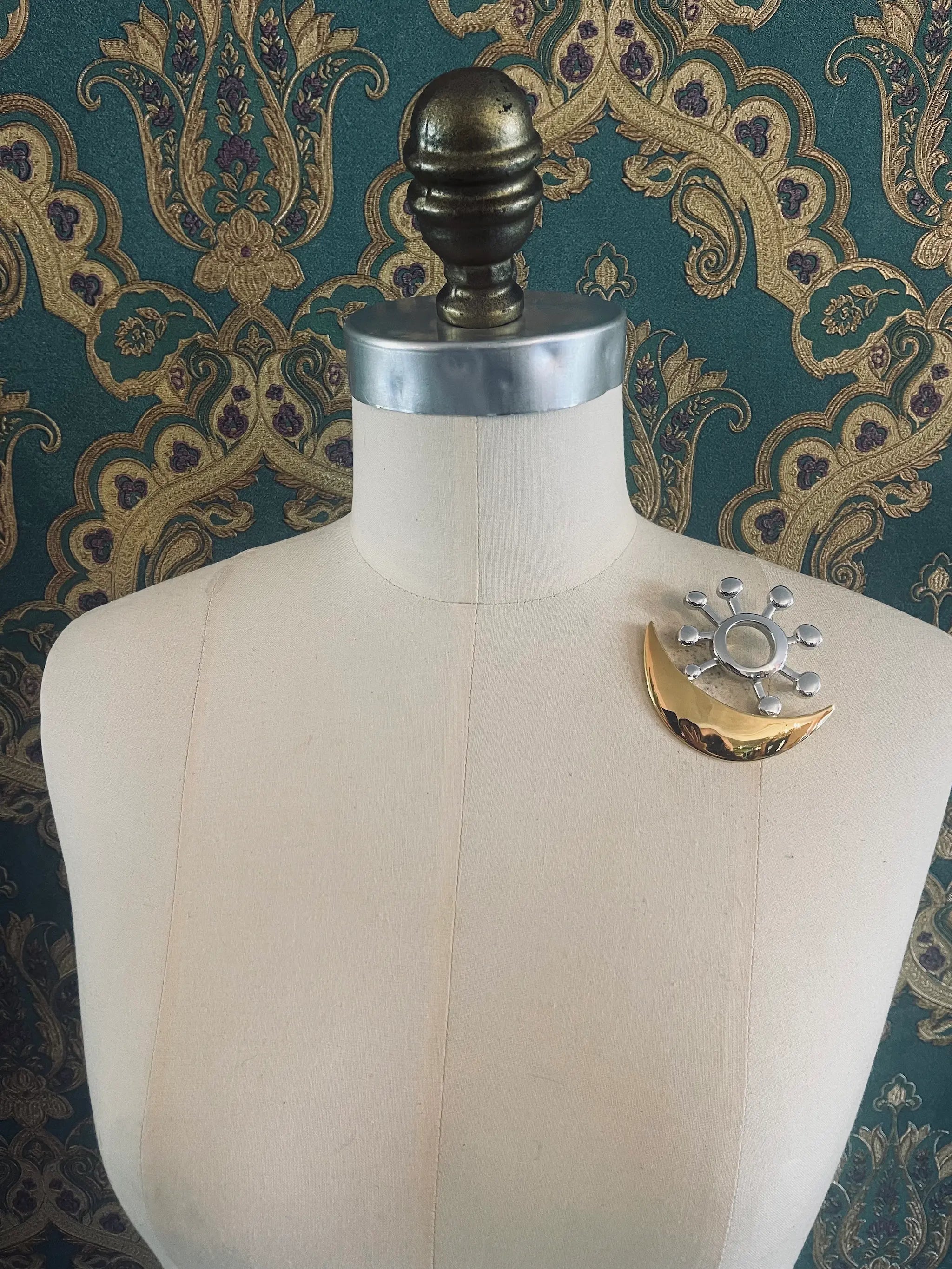 Broochella Brooches with meaning love and harmony designer brooch in gold and silver metal. Pictured on a mannequin. The background is green and gold damask wallpaper