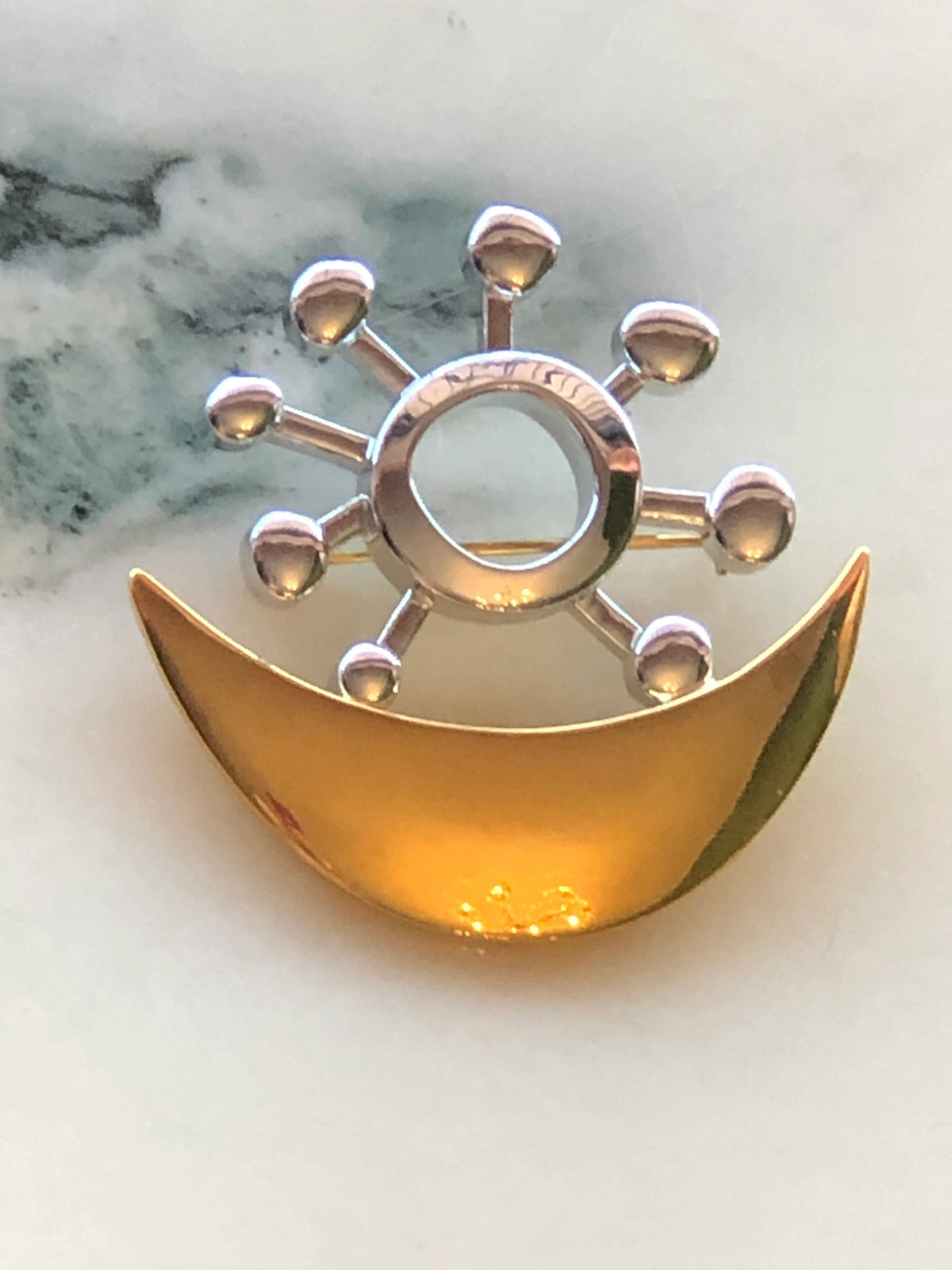 Broochella Brooches with Meaning Love and Harmony Brooch Pin featuring a polished gold-tone crescent with a silver-tone circular sun motif on a cream marble background.