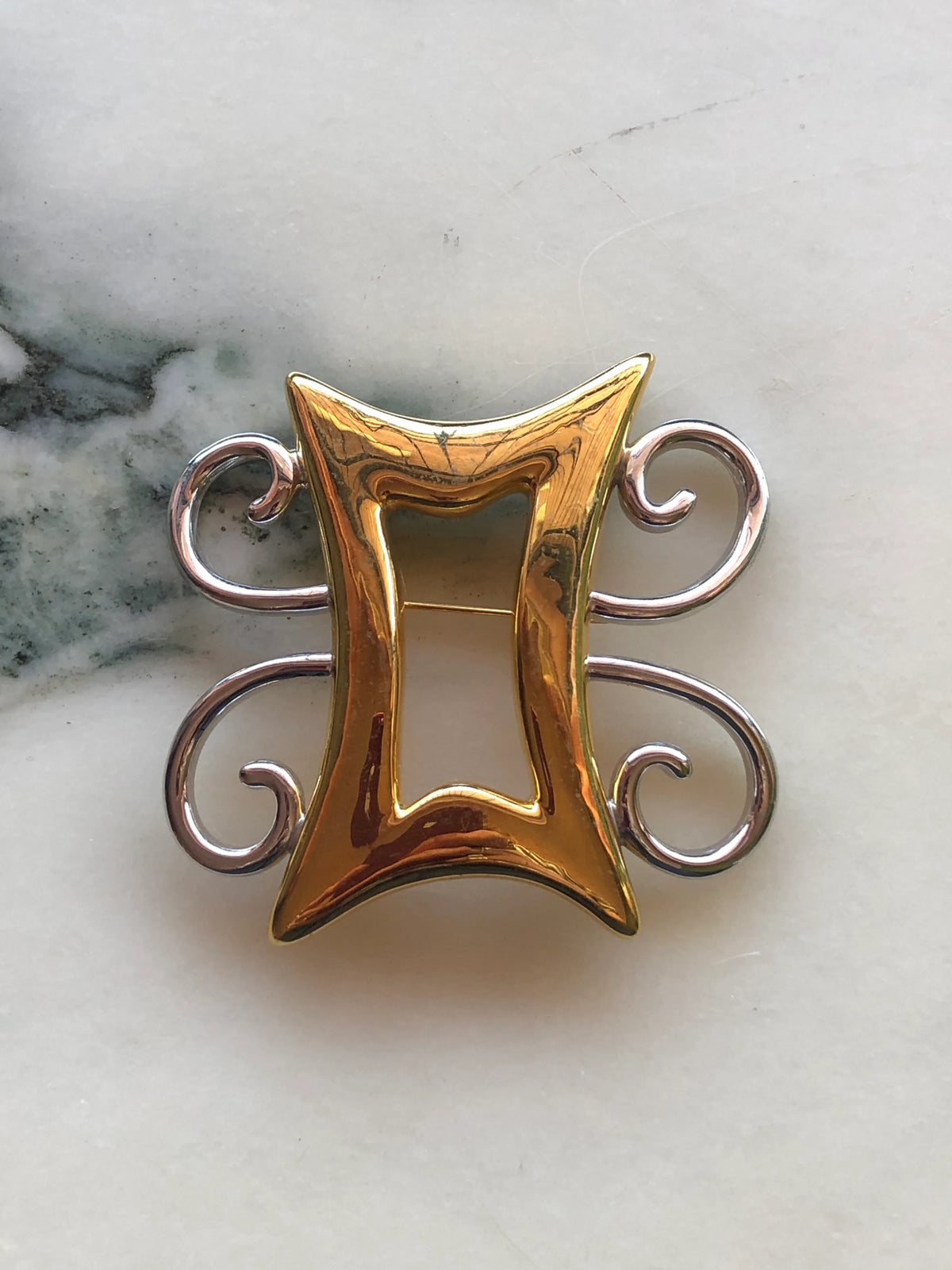 Broochella Brooches with Meaning – Strong & Humble brooch in polished gold and silver-tone metal, featuring an open geometric centre with elegant curled detailing.