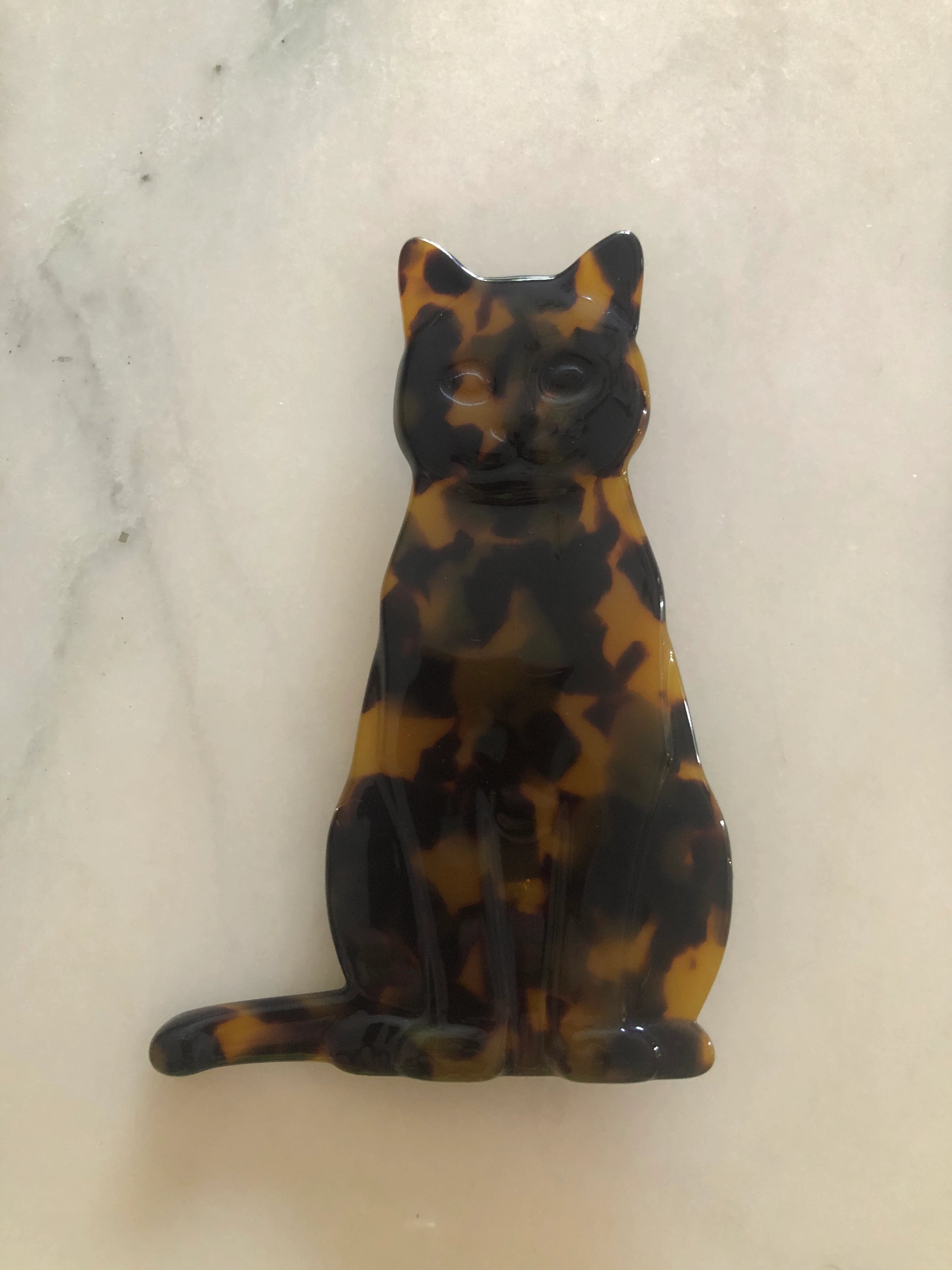 A Broochella Animalia glossy tortoiseshell cat-shaped brooch or pin, featuring a black and amber-brown mottled pattern, displayed on a light marble surface.
