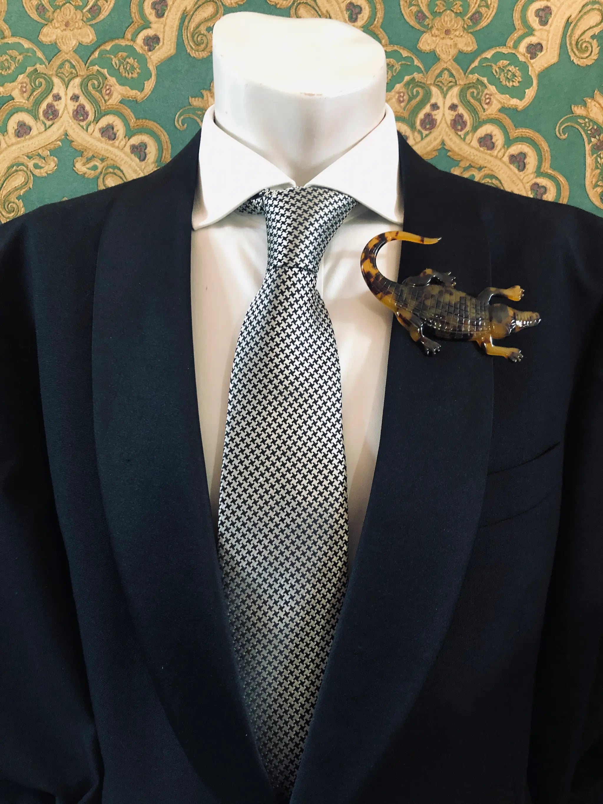 Broochella Men's brown Animalia tortoiseshell crocodile shaped brooch. It is made from Premium Italian cellulose tortoiseshell acetate. It is presented on a men's mannequin that is dressed in a black suit. The background is green and gold damask wallpaper. 