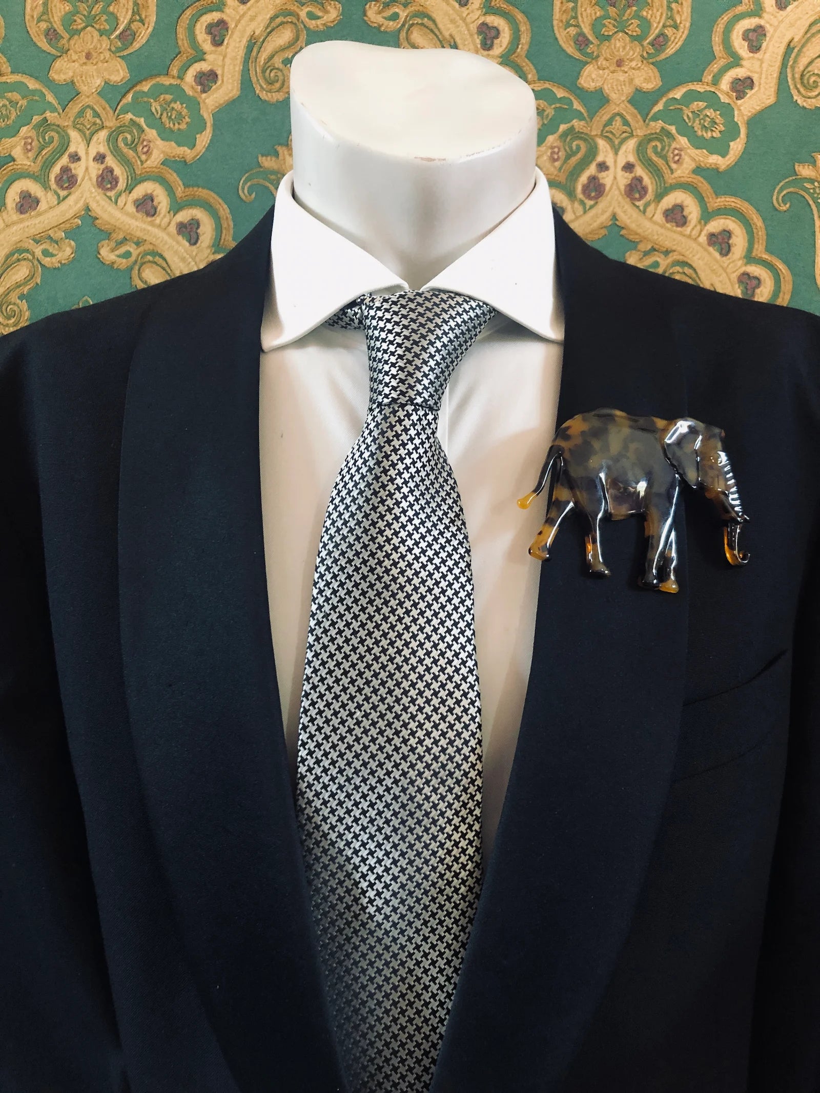 Broochella Men's brown Animalia tortoiseshell giraffe shaped brooch. It is made from Premium Italian cellulose tortoiseshell acetate. It is presented on a men's mannequin that is dressed in a black suit. The background is green and gold damask wallpaper. 