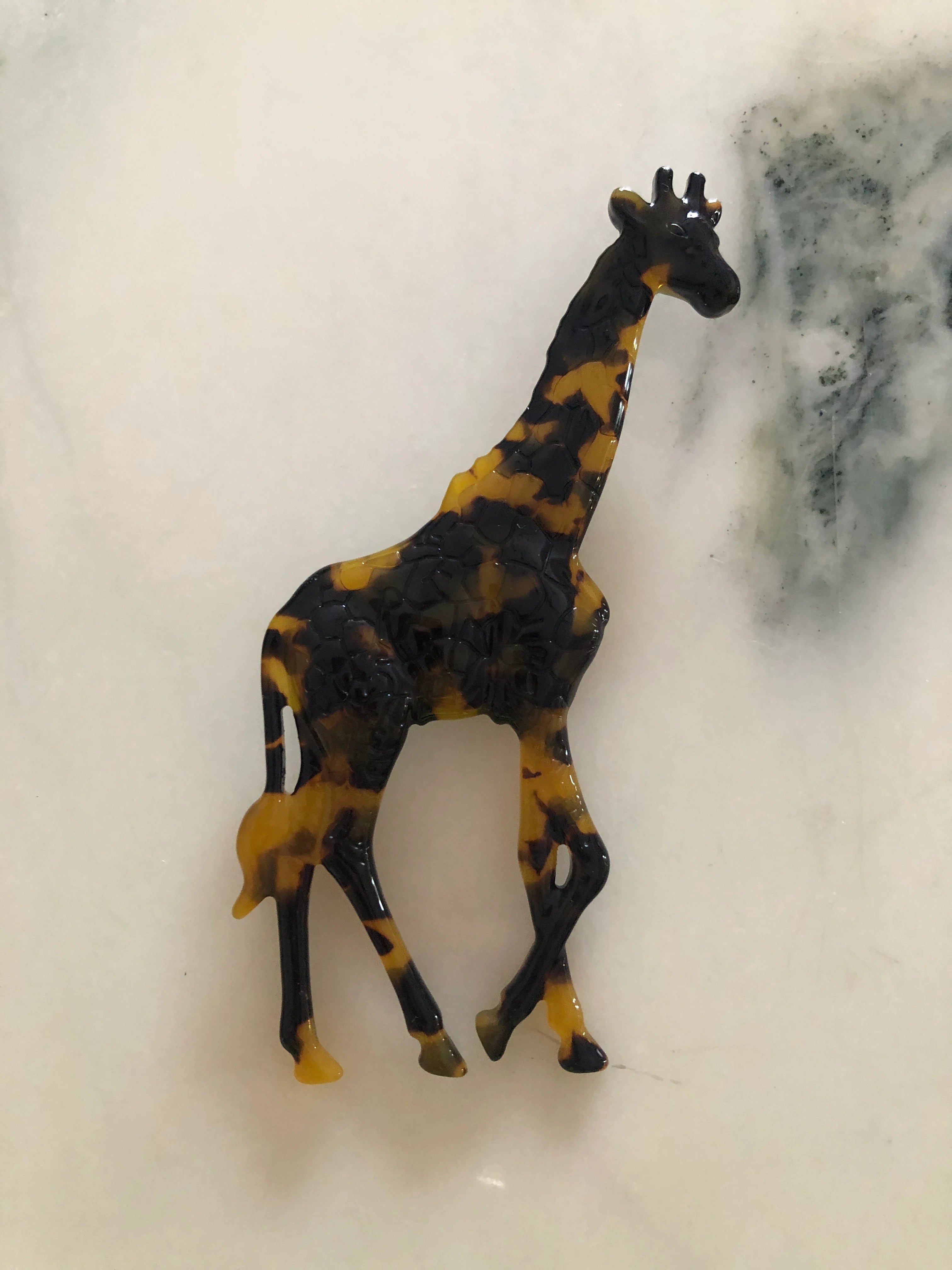 A Broochella Animalia Collection standing giraffe brooch in brown, black and beige tortoiseshell Italian cellulose acetate with sculpted spot patterning measuring 12cm x 7cm.  The background is white marble.