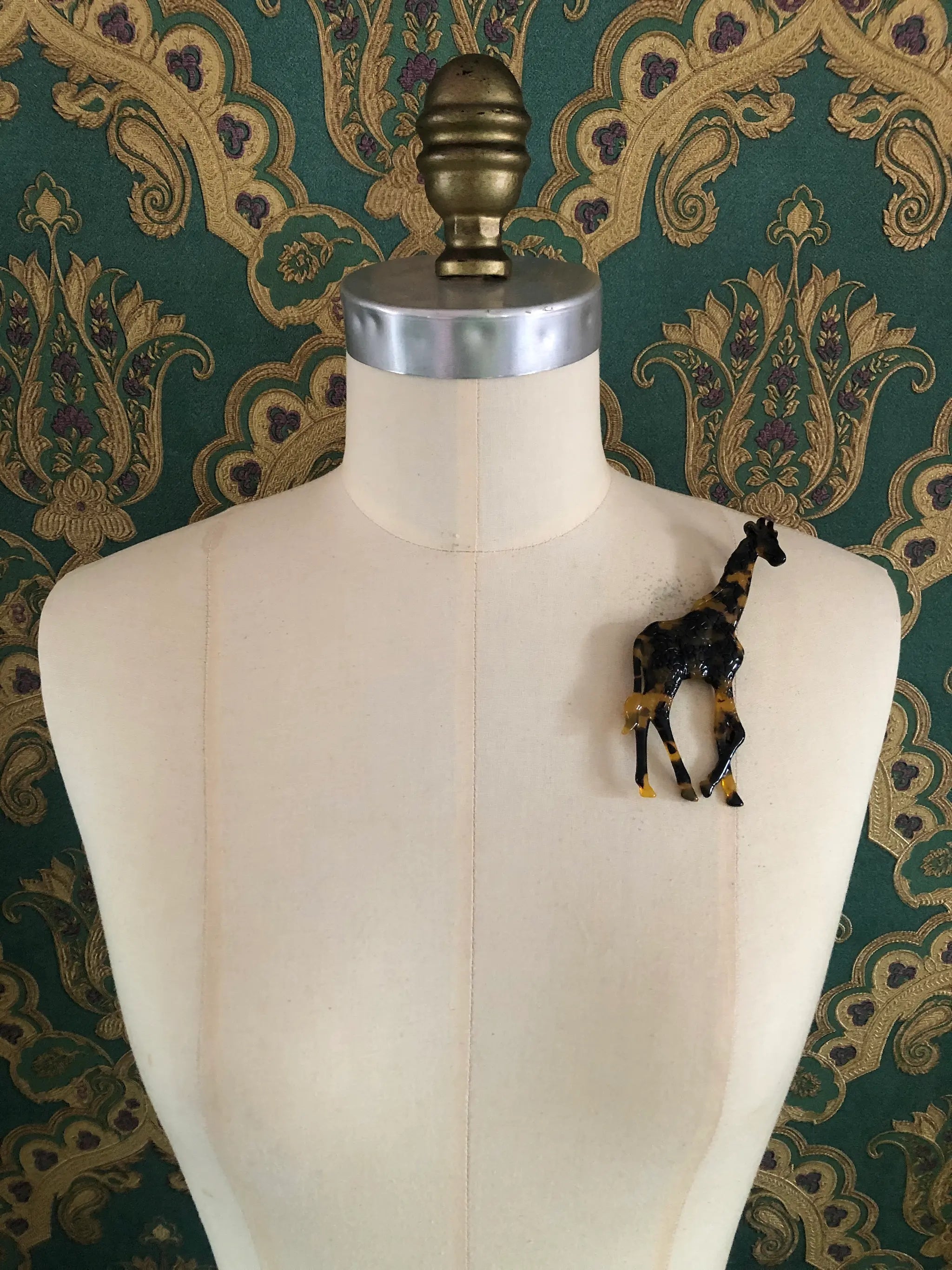 A Broochella Animalia Collection standing giraffe brooch in brown, black and beige tortoiseshell Italian cellulose acetate with sculpted spot patterning measuring 12cm x 7cm. It is pinned on a mannequin. The background is green and gold damask wallpaper. 