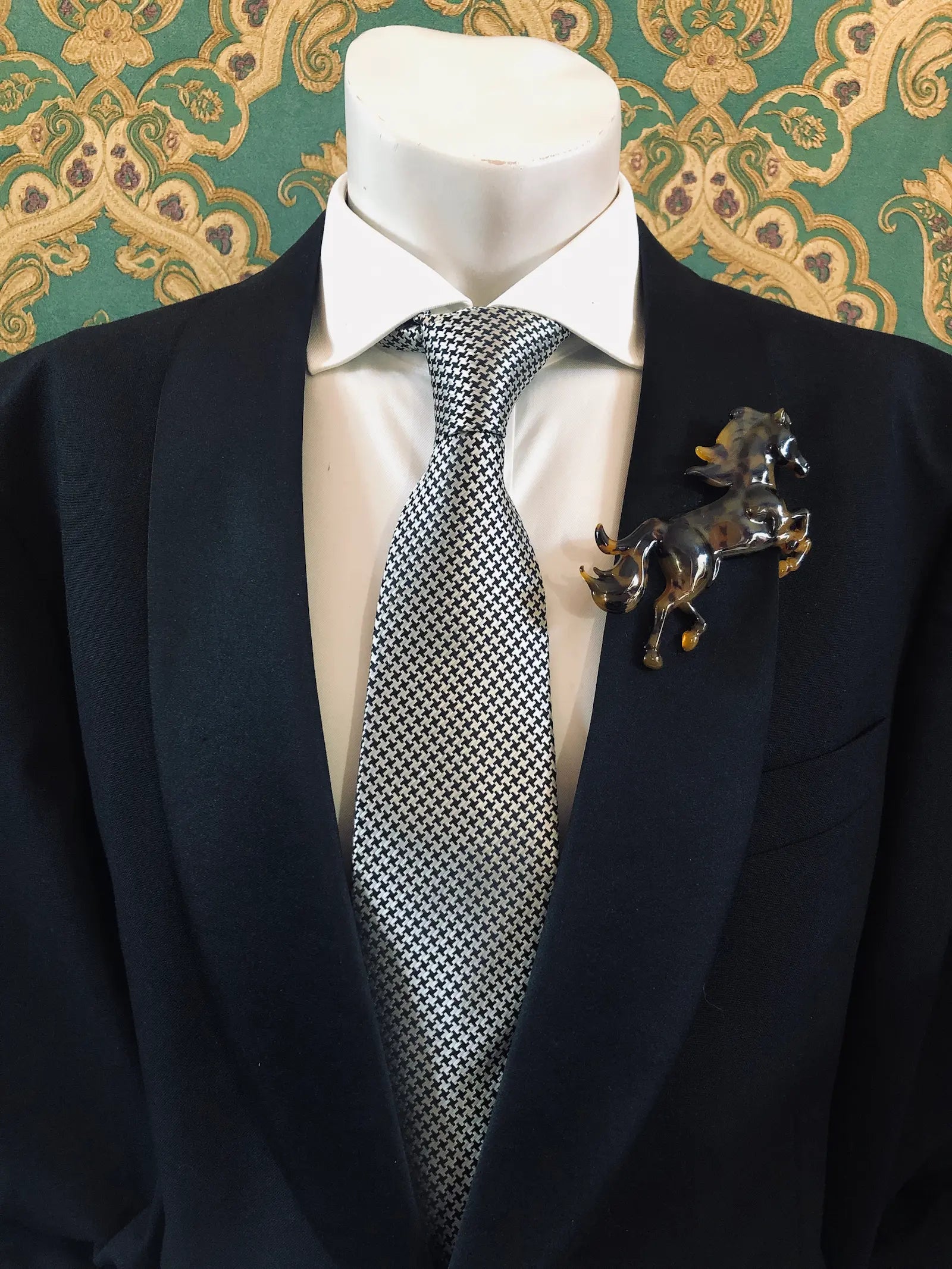 Broochella Men's Animalia  brown and black tortoiseshell figural horse brooch. It is pinned to a men's mannequin that is dressed in a black suit. The background is green and gold damask wallpaper. 