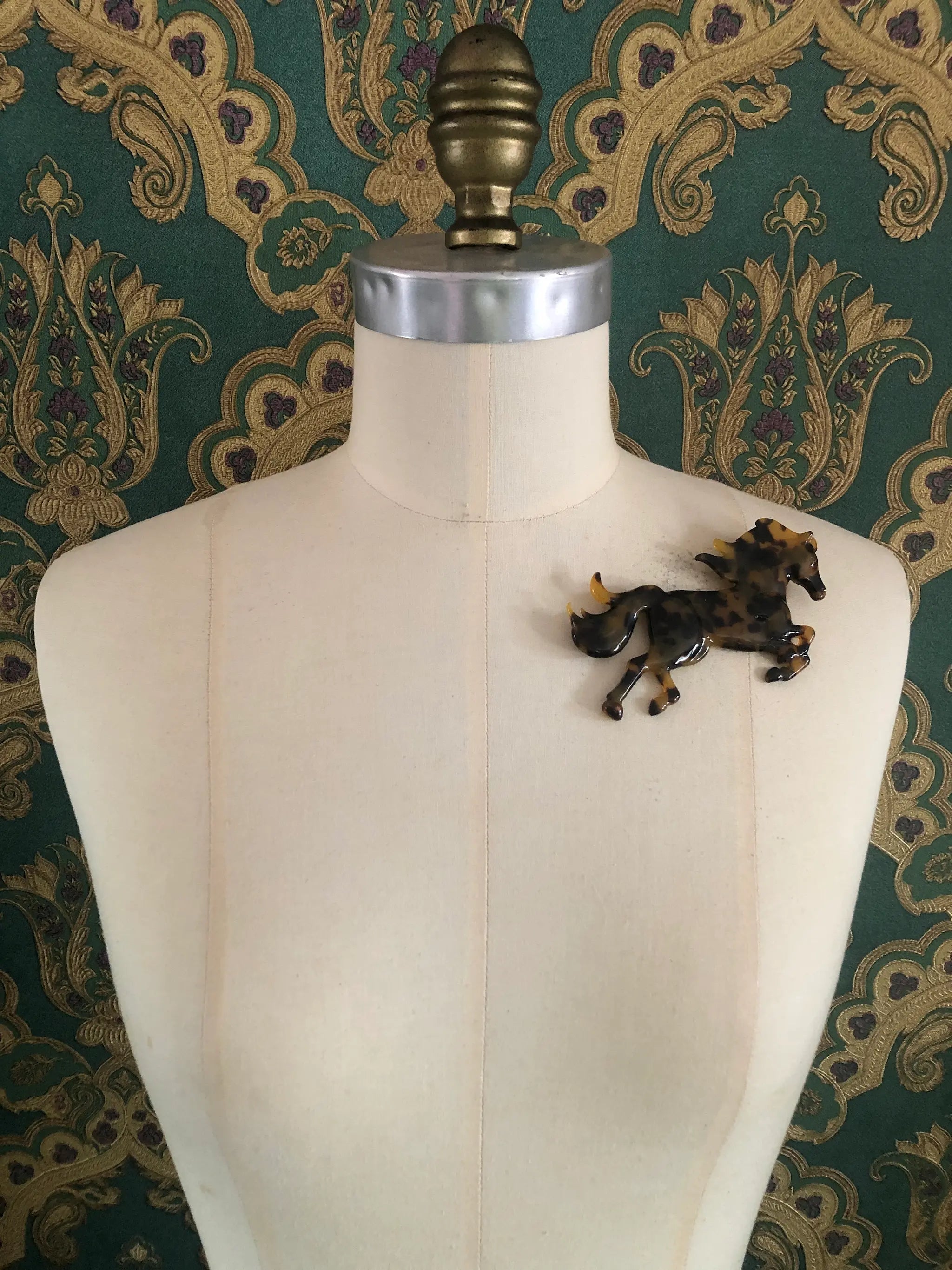 Broochella Animalia Collection black and brown tortoiseshell horse brooch. It is made from premium, Italian cellulose acetate. The brooch is pinned to a mannequin. The background is green and gold damask wallpaper. 