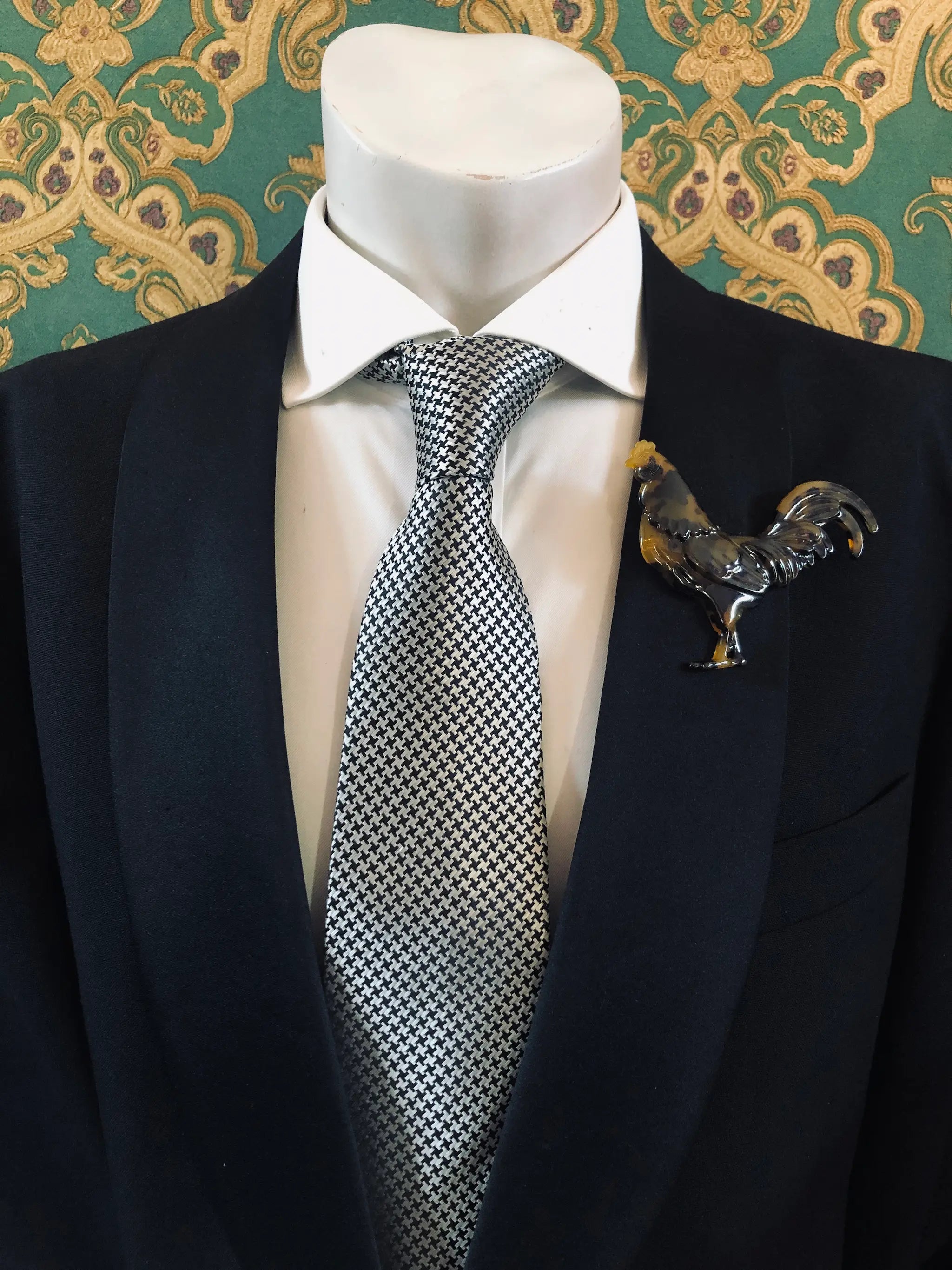A Broochella Animalia Men's tortoiseshell rooster brooch displayed on a white mannequin torso, featuring a black  suit jacket, a white dress shirt, a silver and black checkered tie against a green and gold patterned wallpaper background.