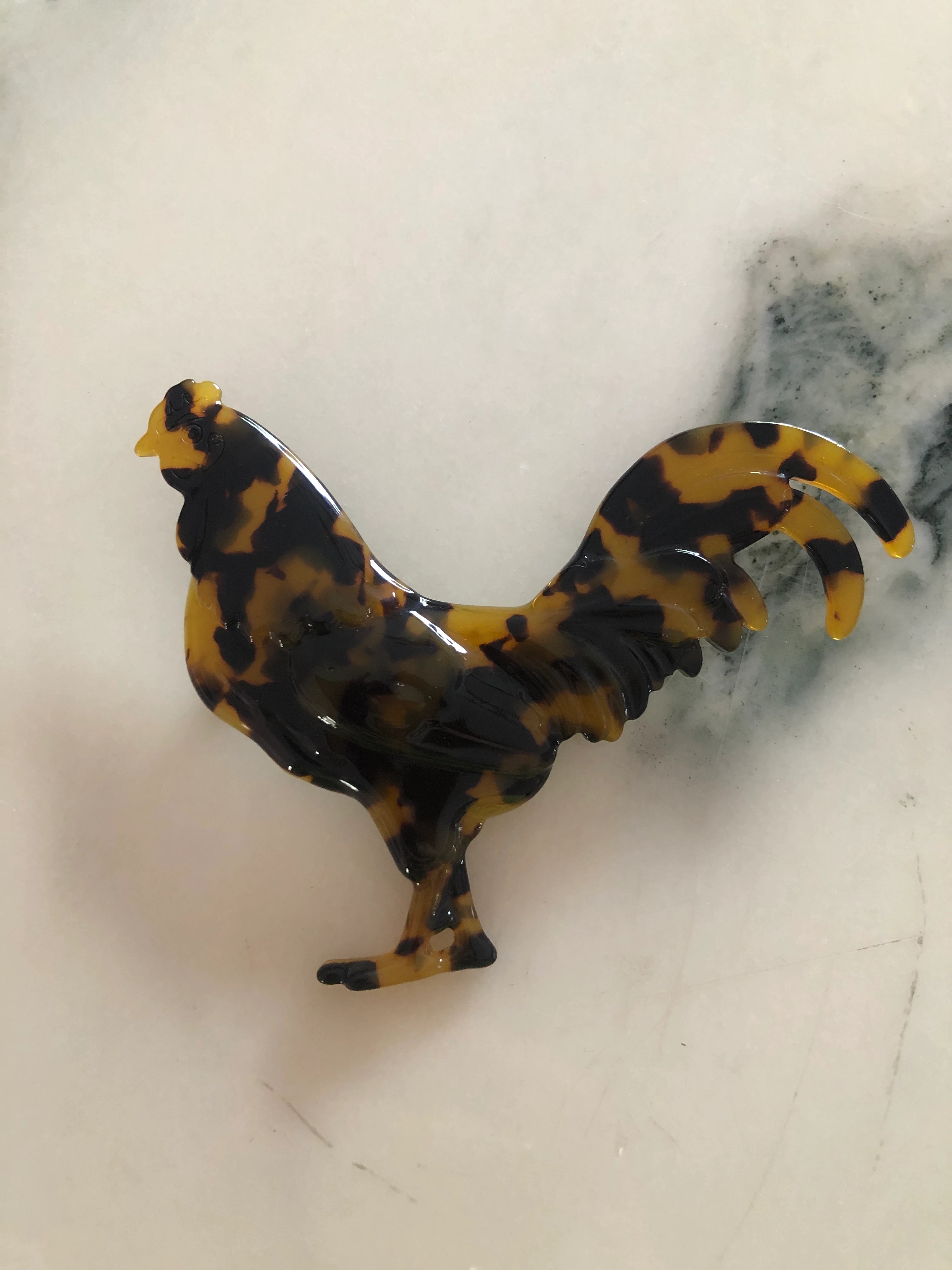A Broochella Animalia Collection rooster-shaped brooch made of glossy black and amber-yellow marbled, tortoiseshell,  Italian cellulose acetate.  shown on a light off-white surface.