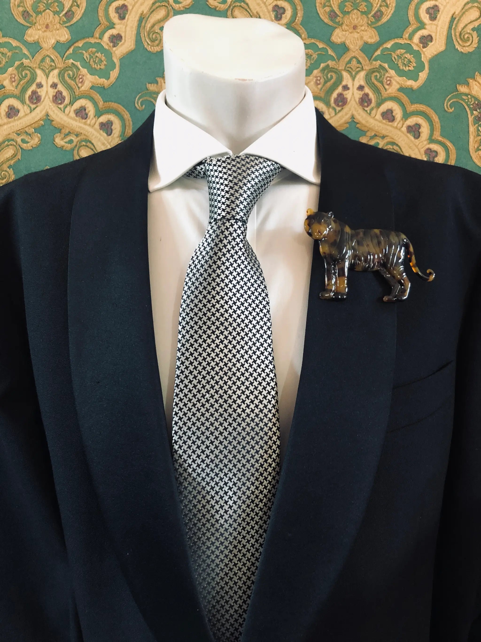 Broochella Men's brown Animalia tortoiseshell tiger shaped brooch. It is made from Premium Italian cellulose tortoiseshell acetate. It is presented on a men's mannequin that is dressed in a black suit. The background is green and gold damask wallpaper. 