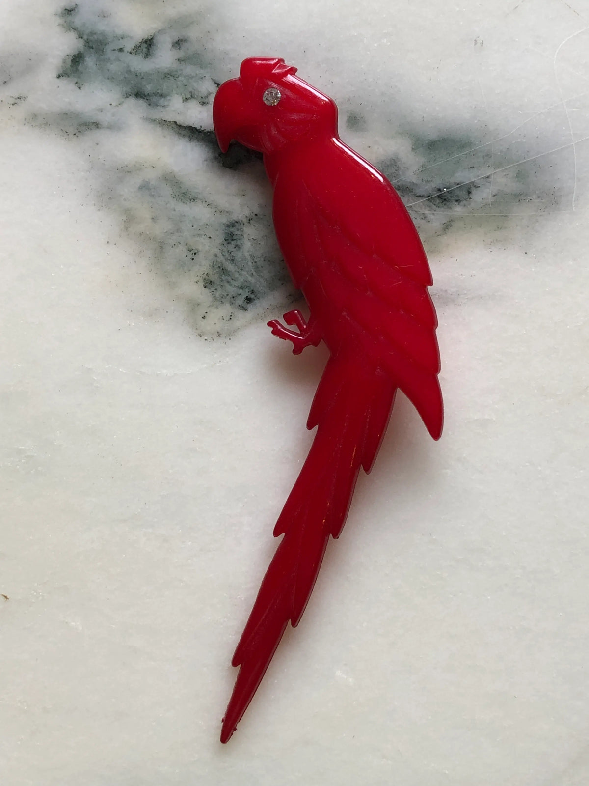 Broochella vintage Buch + Deichmann Scarlet Macaw brooch in vivid red with a clear crystal eye and long sculptural tail, made in Copenhagen, Denmark and measuring approximately 14 × 6 cm. Displayed on white marble