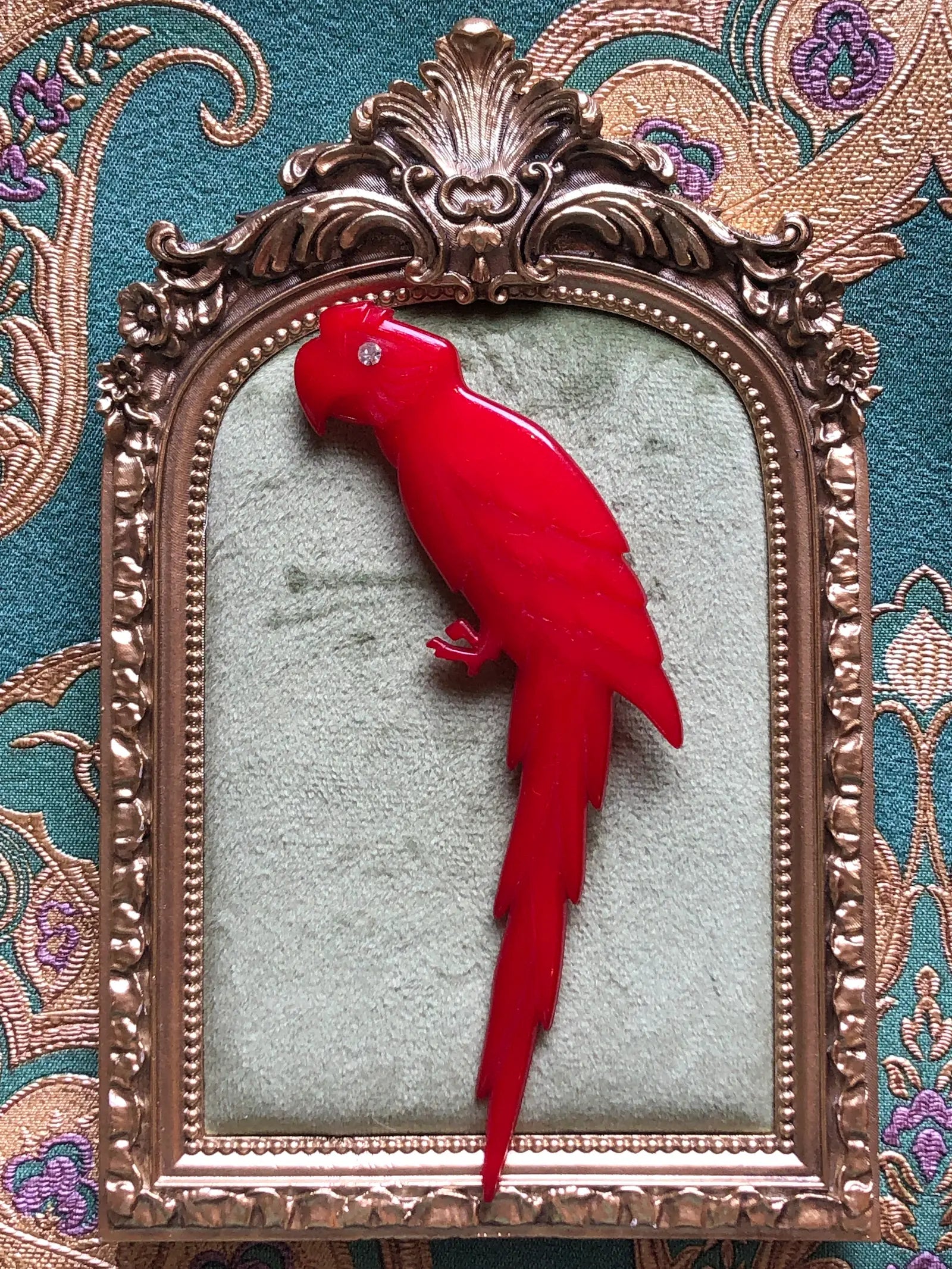 Broochella vintage Buch + Deichmann Scarlet Macaw brooch in vivid red with a clear crystal eye and long sculptural tail, made in Copenhagen, Denmark and measuring approximately 14 × 6 cm. Displayed on green velvet in an ornate gold-tone frame.