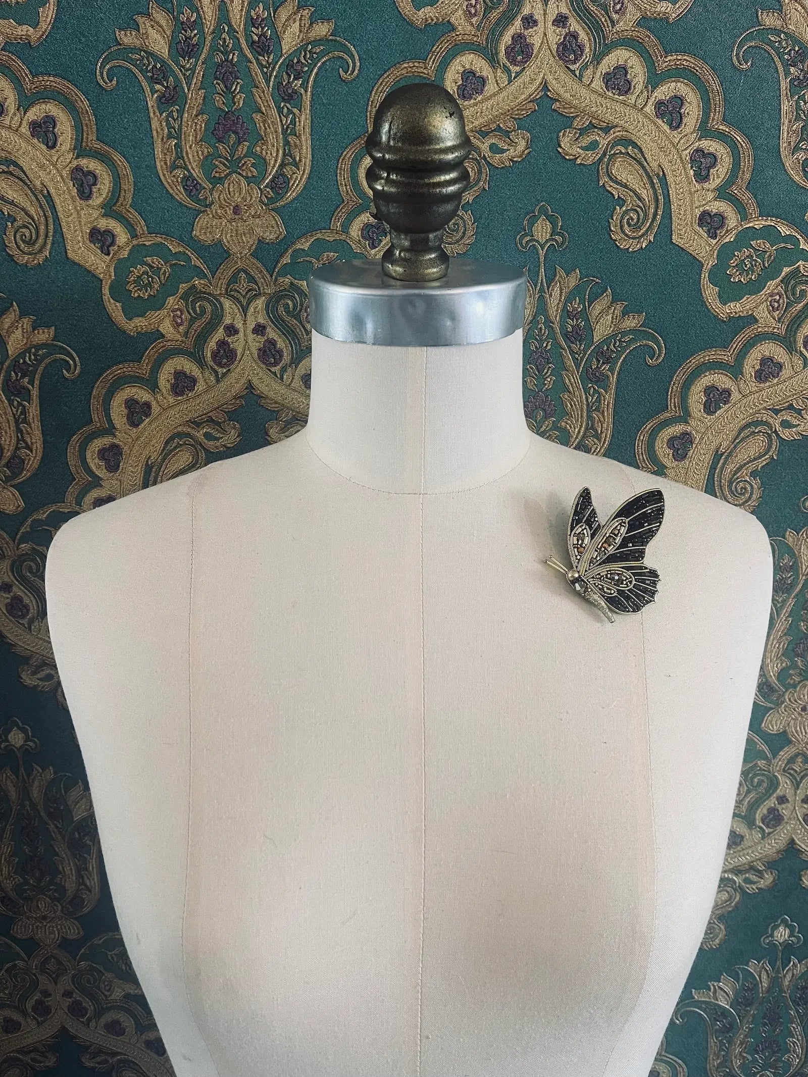 Broochella hand beaded black and gold butterfly brooch. Pictured on a mannequin. The background is green and gold damask wallpaper