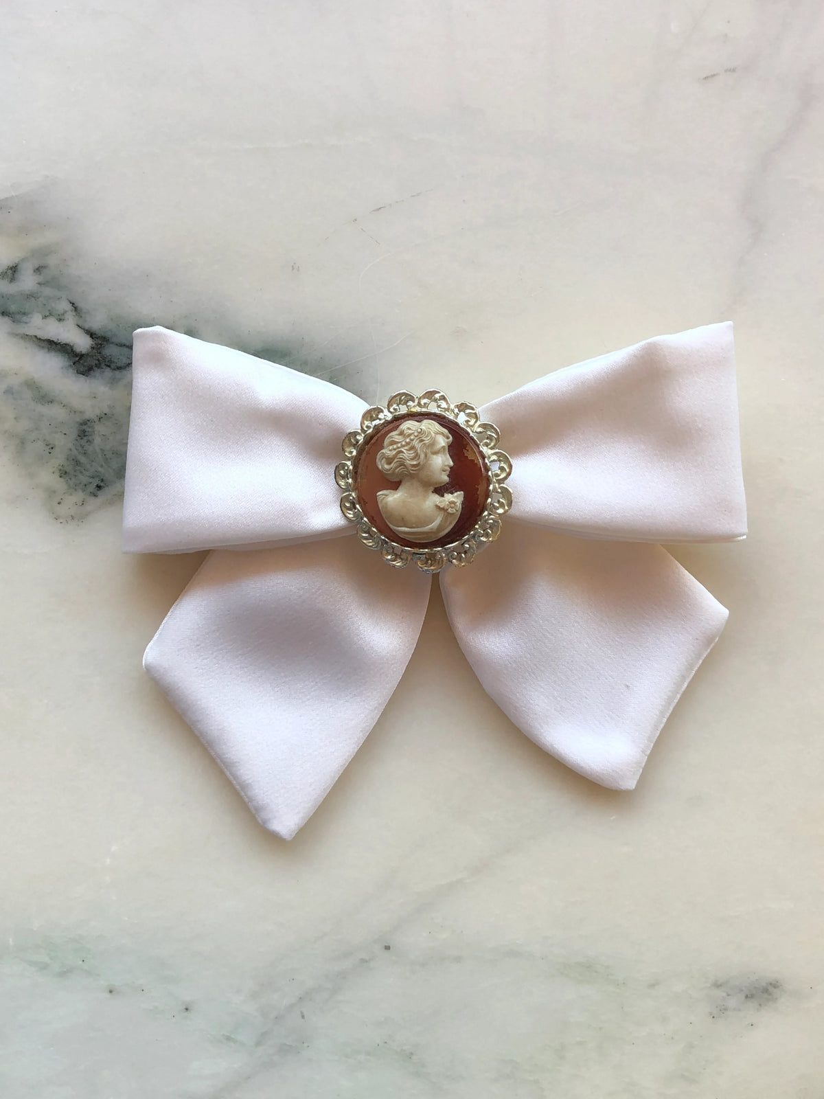 Broochella Camée Ivoire Ivory Cameo Bow Brooch featuring an ivory satin bow with a cream portrait cameo on a warm brown ground in an ornate silver-tone setting, photographed on white marble with soft grey-green veining.