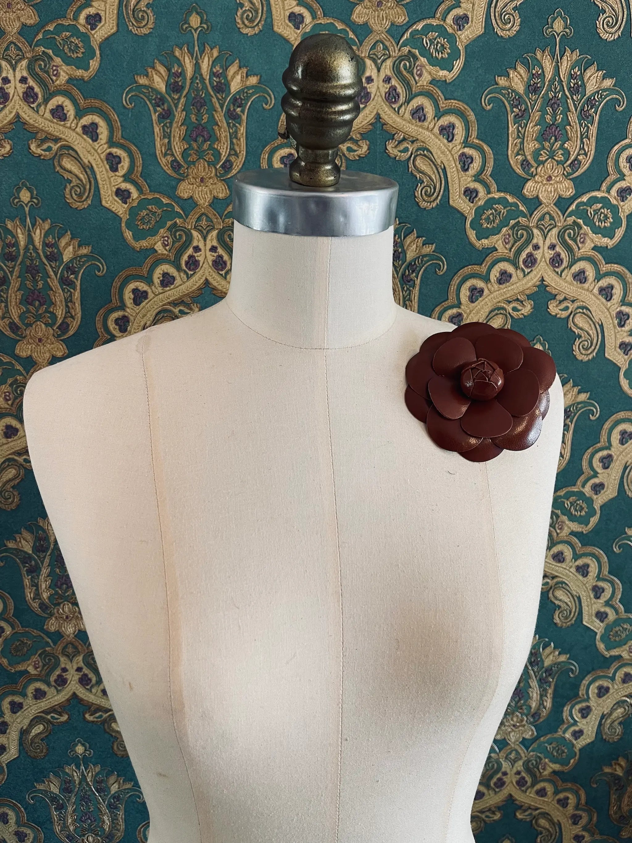Broochella leather handmade designer camelia flower brooch. Styled on a mannequin. The background is green and gold damask wallpaper