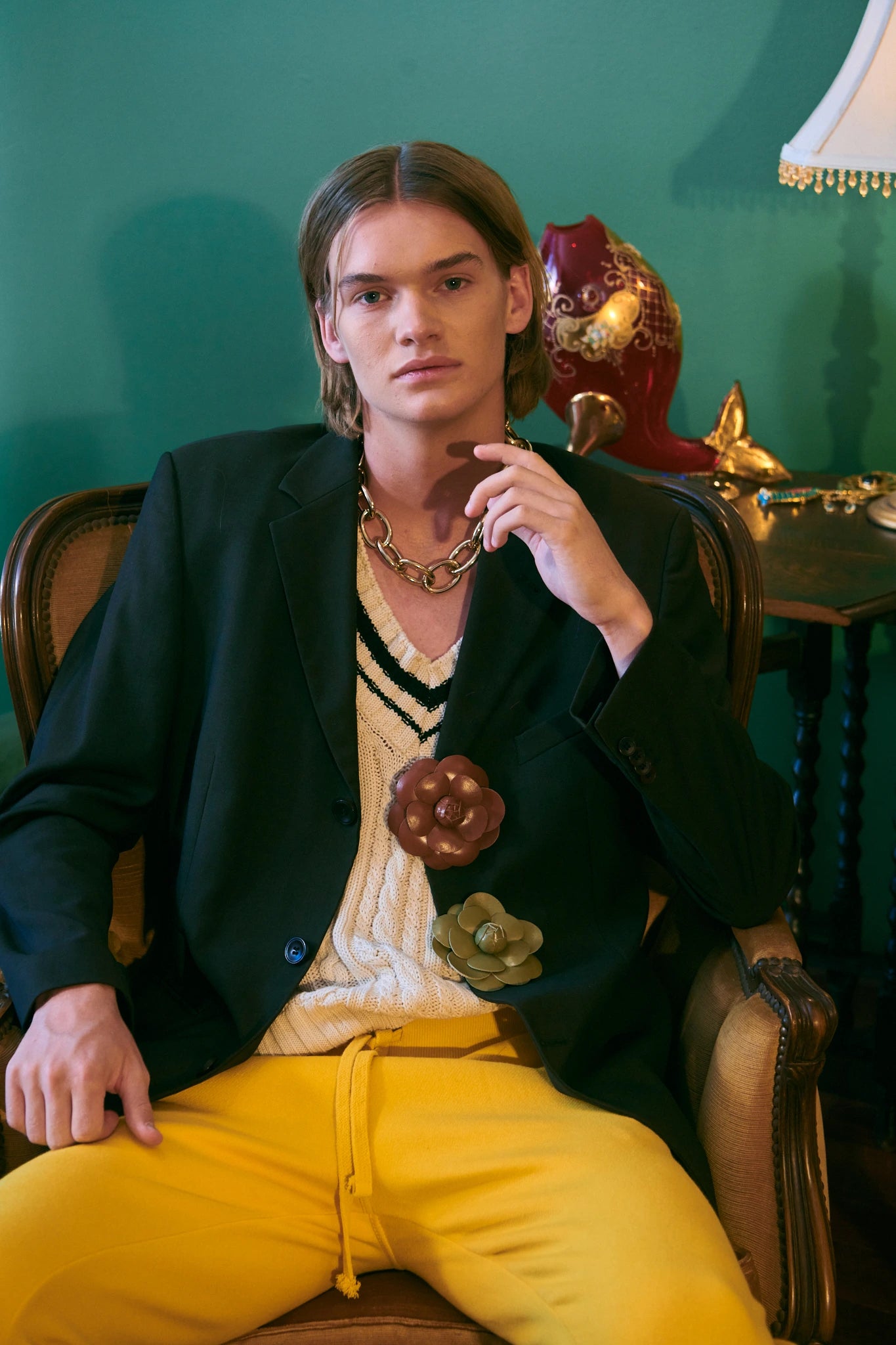 A model wearing a black blazer with camelia leather  flower brooch, a cream knit sweater, and bright yellow pants, seated in a vintage armchair against a dark green wall.