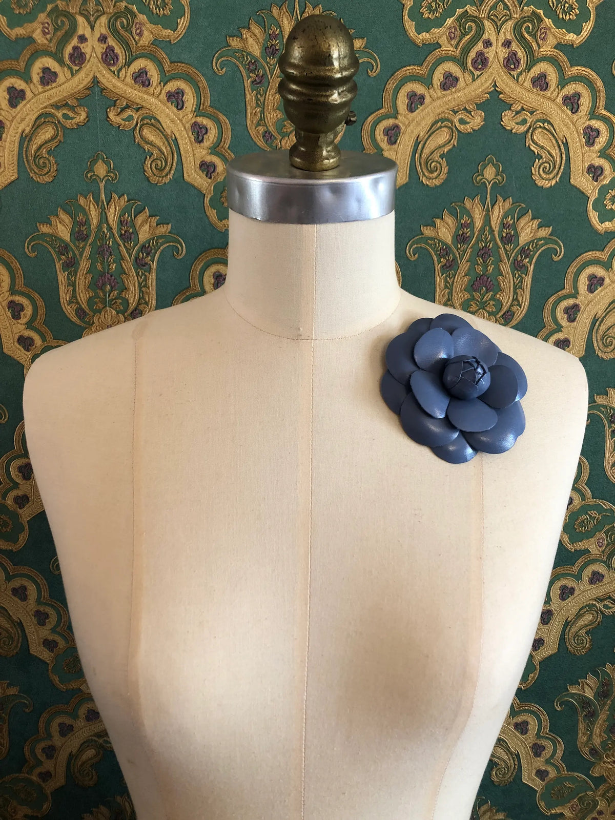 A Broochella Camelia blue leather flower brooch displayed on a cream-colored dress form against a green and gold damask patterned wallpaper background.