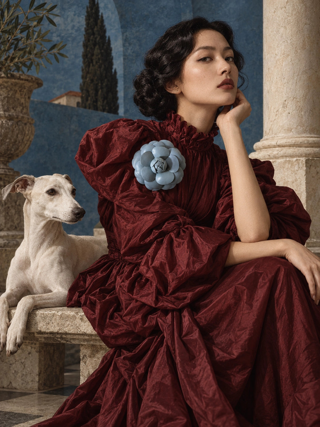 A woman in a voluminous deep burgundy dress with a Broochella  blue camelia flower brooch accent sits on a stone bench next to a white dog, set against a classical painted backdrop with columns and a blue wall.